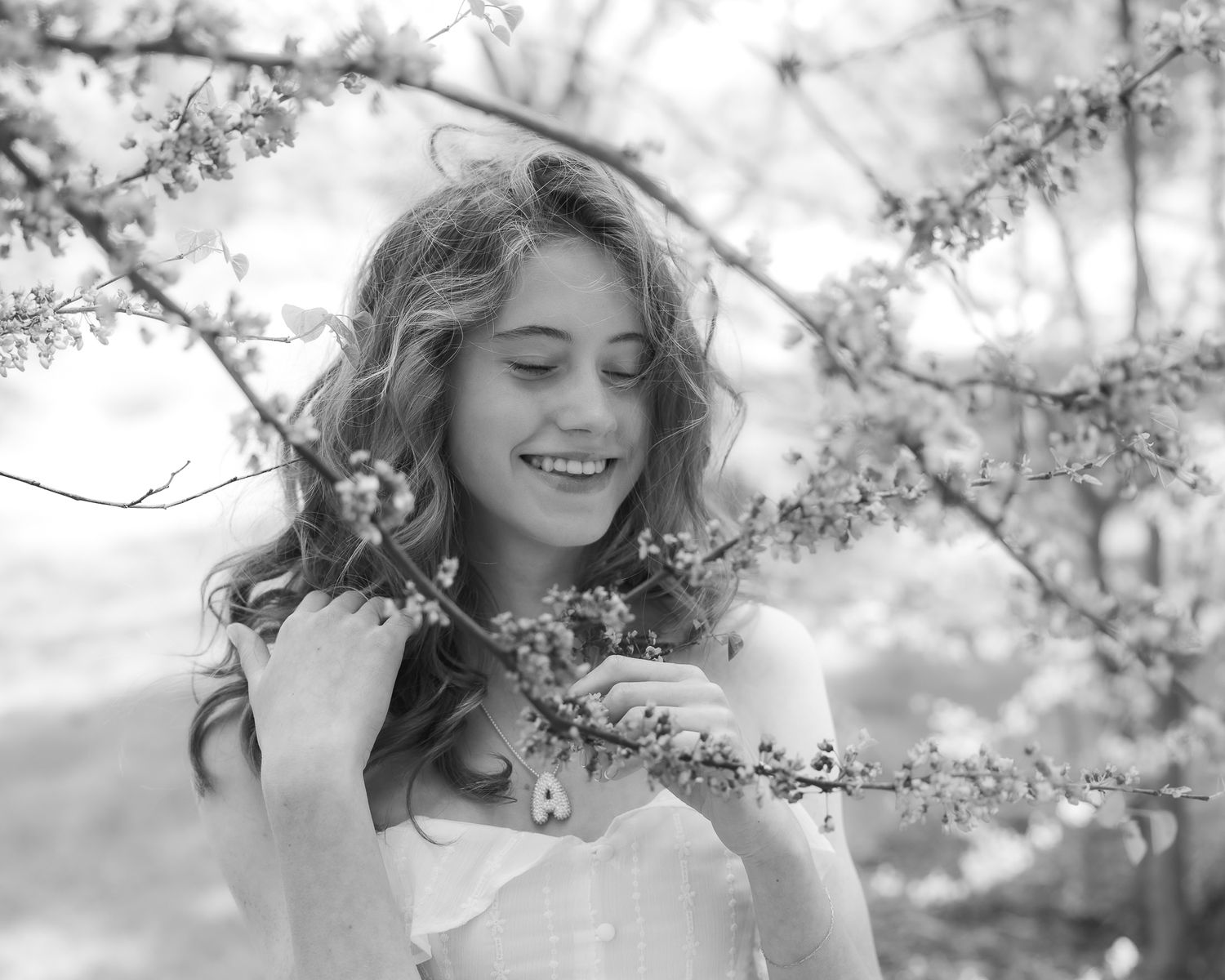 Black and white photo of a woman smiling among spring cherry blossoms wearing a white dress.