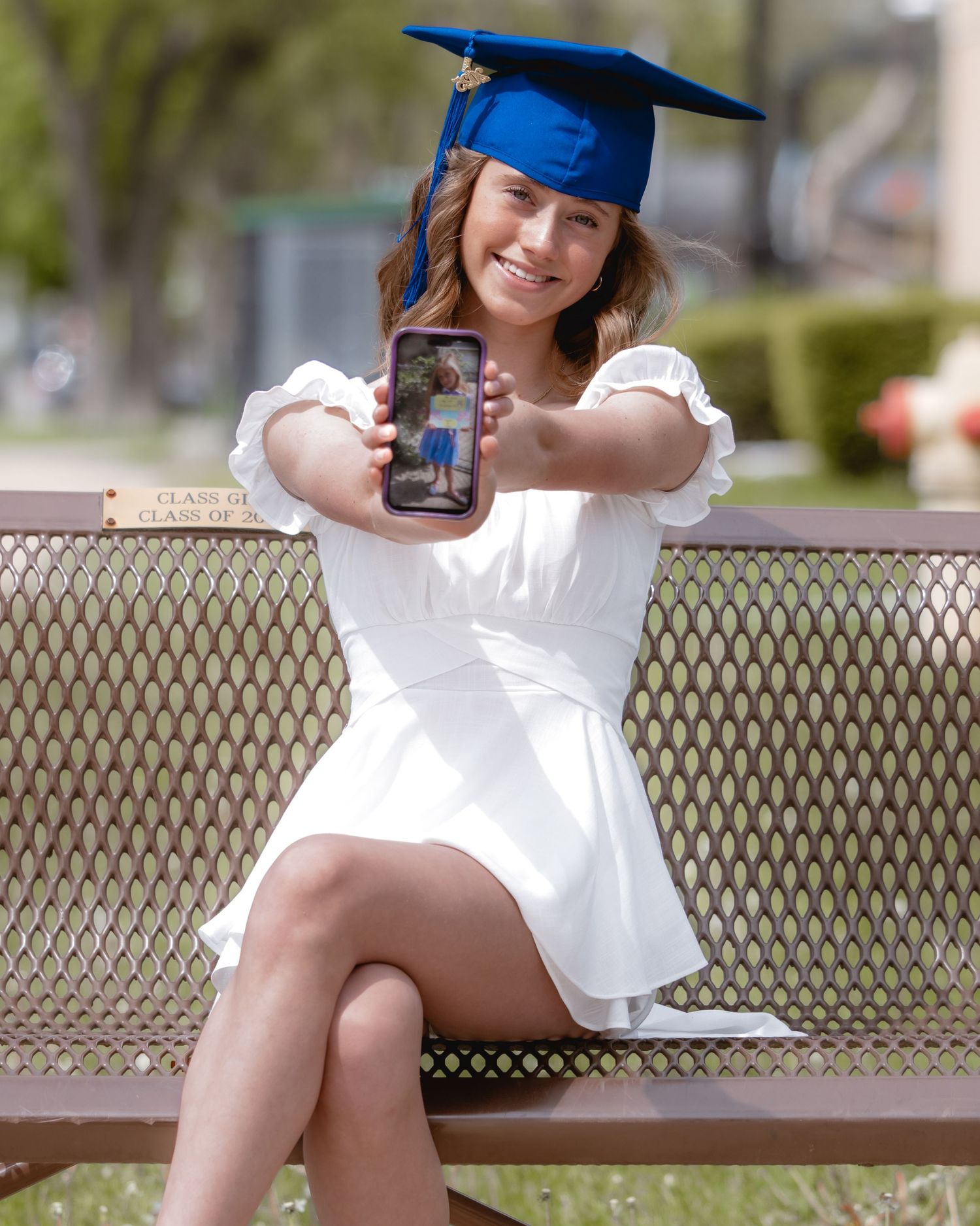 Graduate in white dress and blue cap poses on metal bench outdoors.