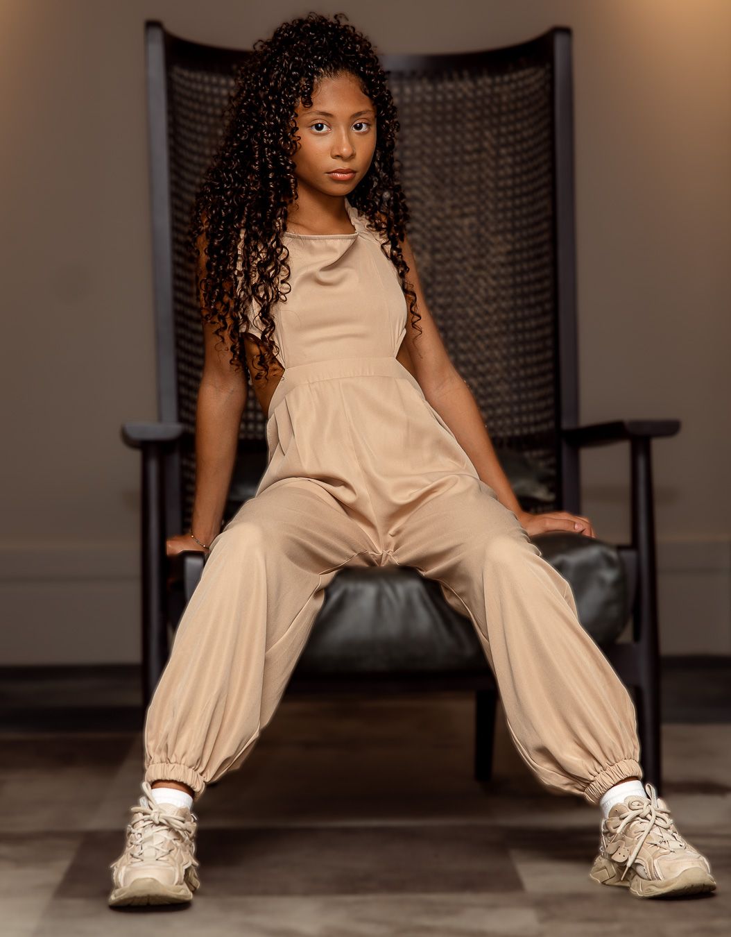 Young girl with curly hair sits confidently in a black chair wearing a beige jumpsuit and white sneakers.