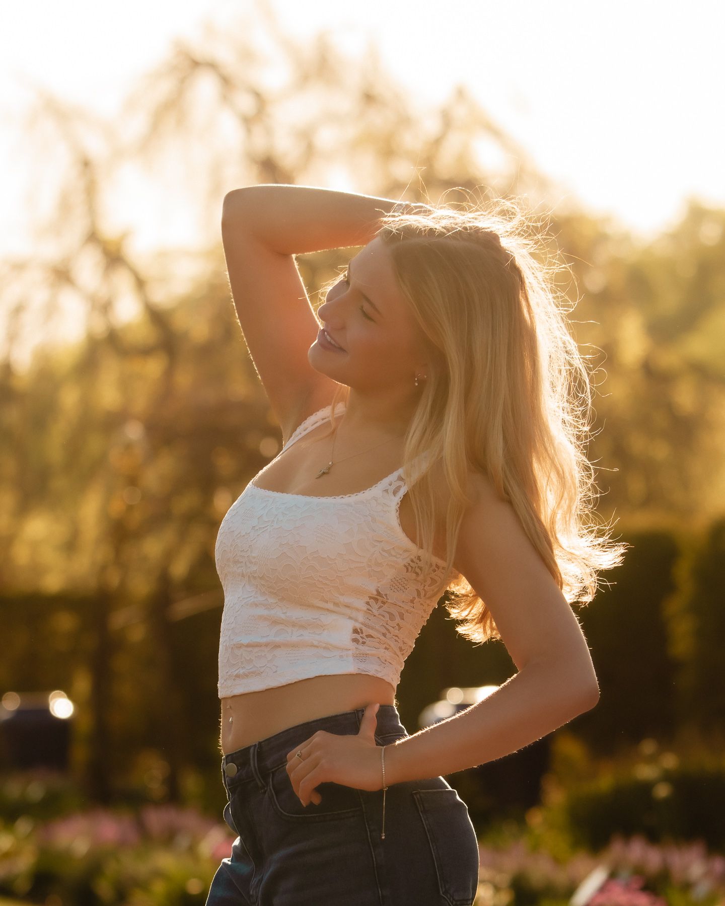 Blonde woman in white crop top and jeans posing outdoors in golden sunset light with trees in background.