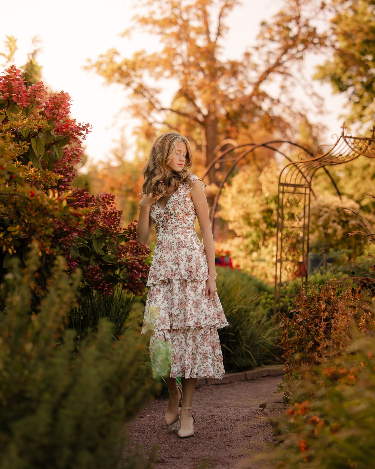 A person in a floral dress walks along a garden path during golden hour with autumn foliage in the background.