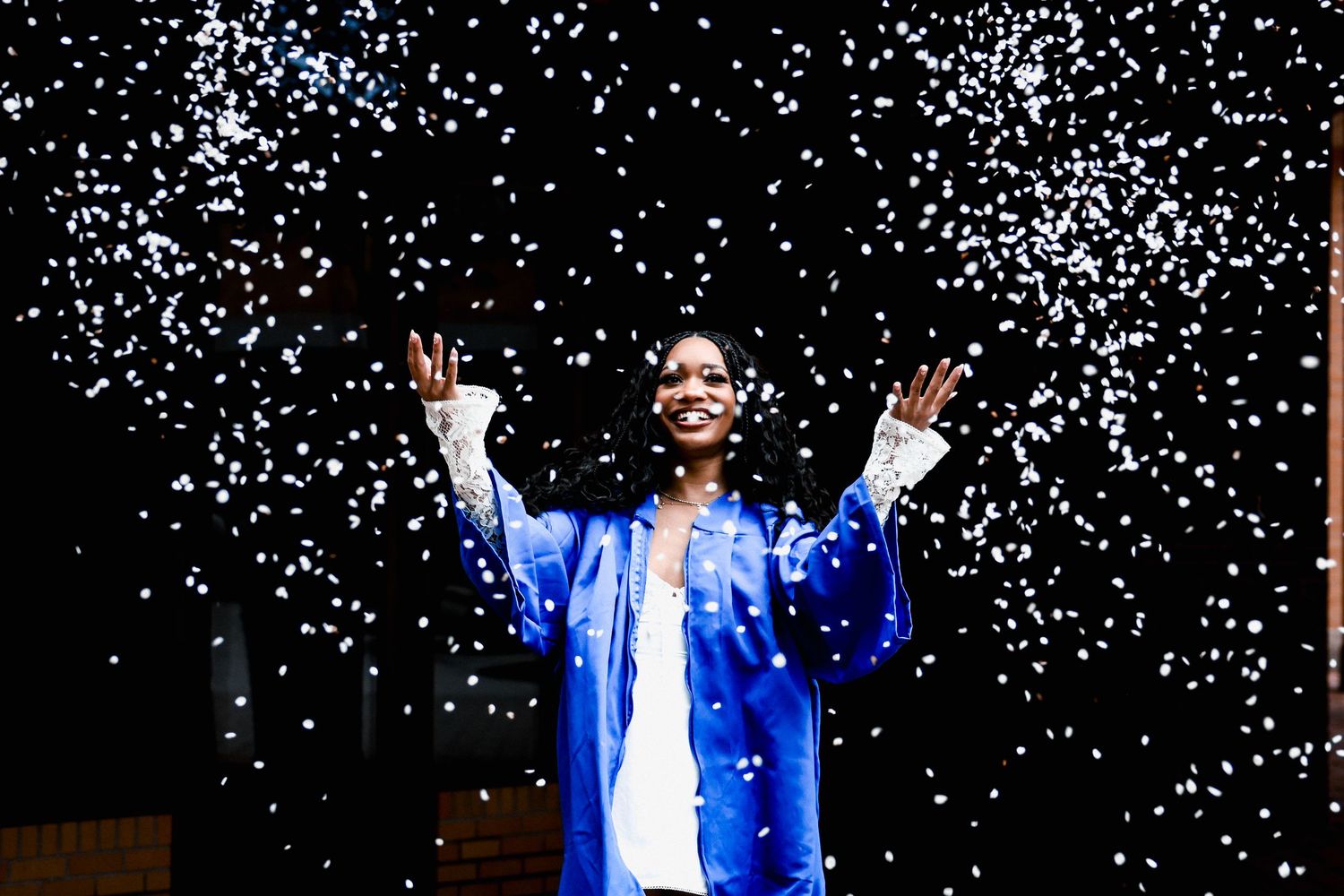 Graduate celebrating with confetti against dark background in blue graduation gown.