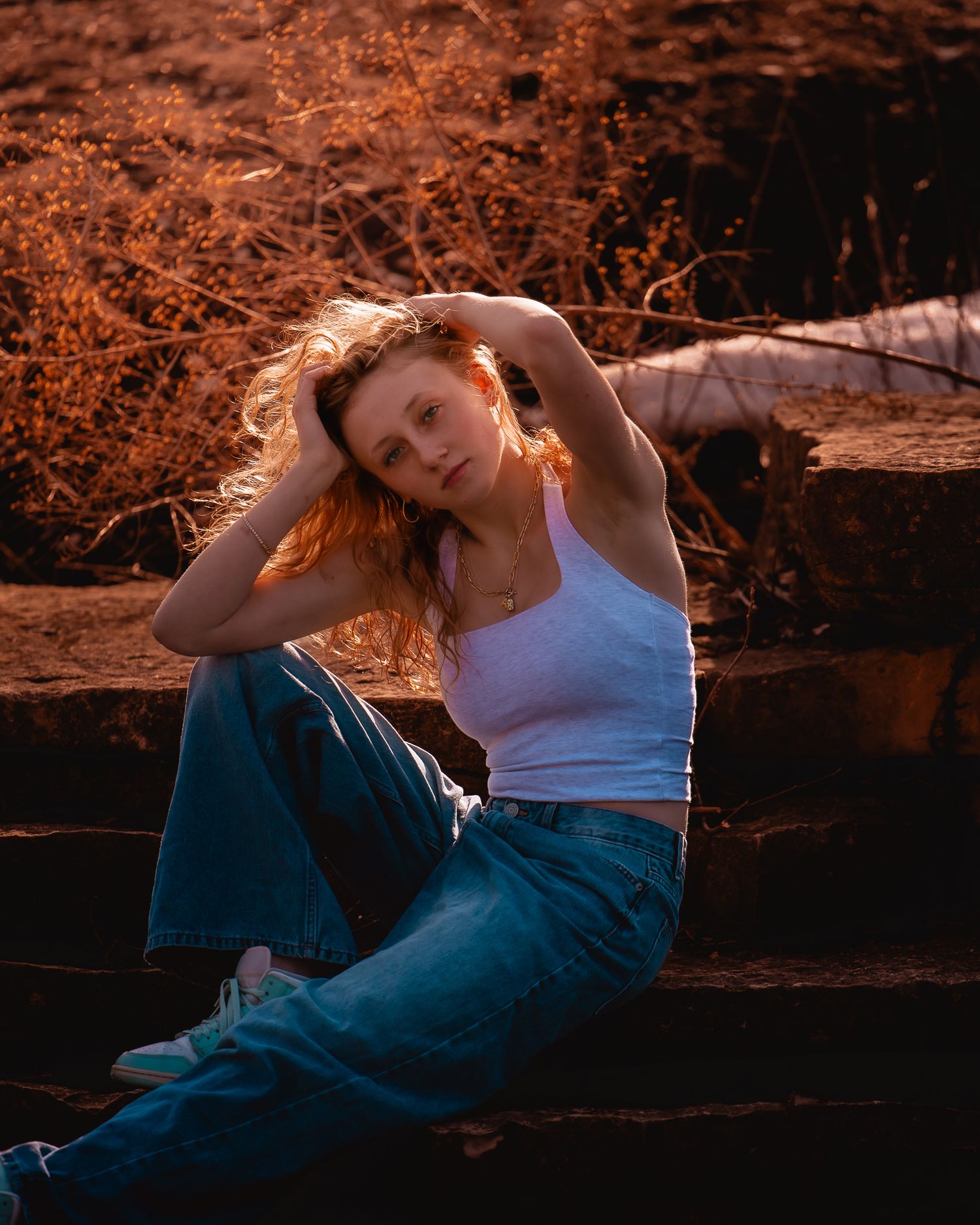 Young woman with wavy red hair sits on stone steps outdoors, wearing a white tank top and wide-leg jeans at golden hour.