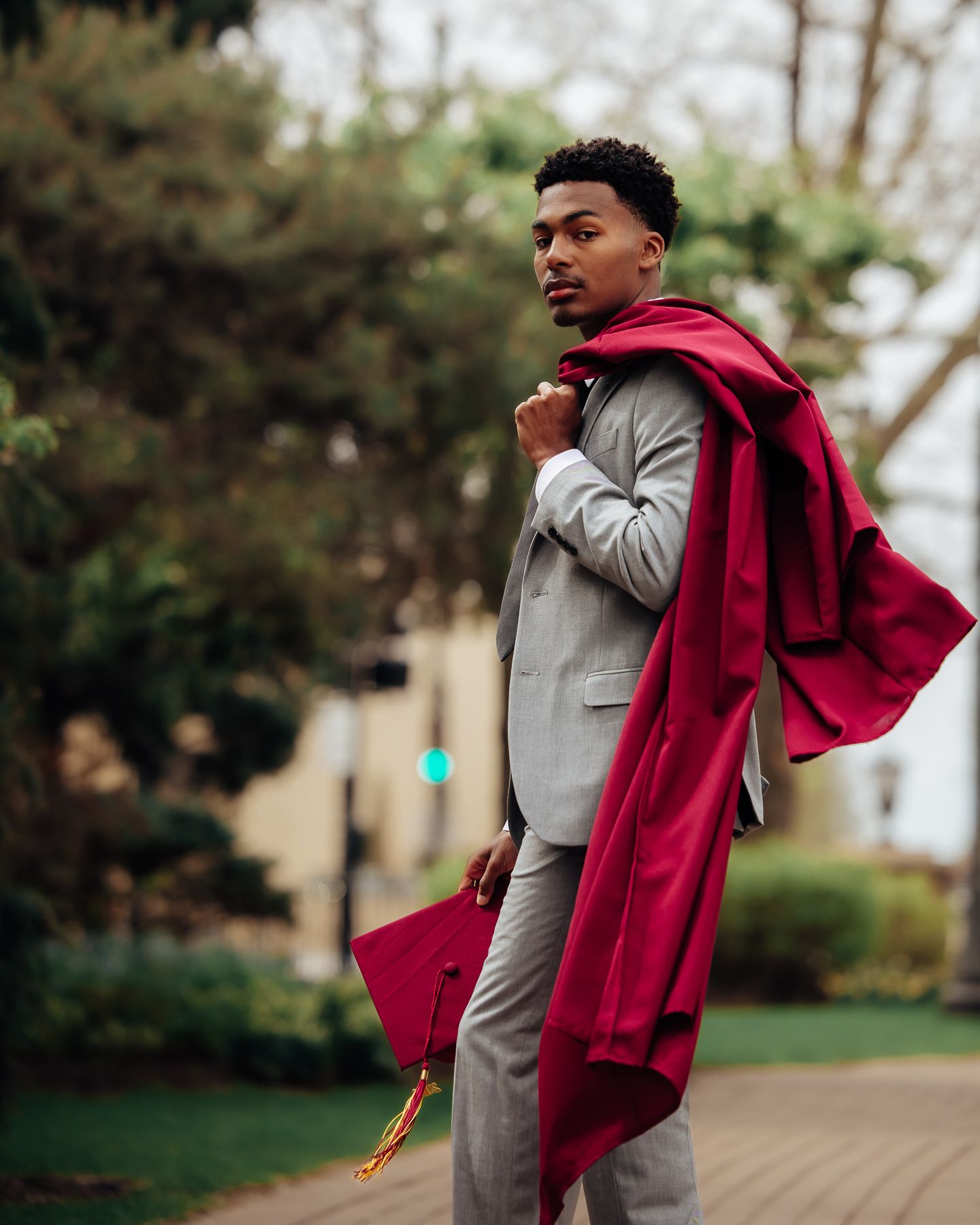 Young man in gray suit wearing red graduation gown and cap poses outdoors on a tree-lined path.