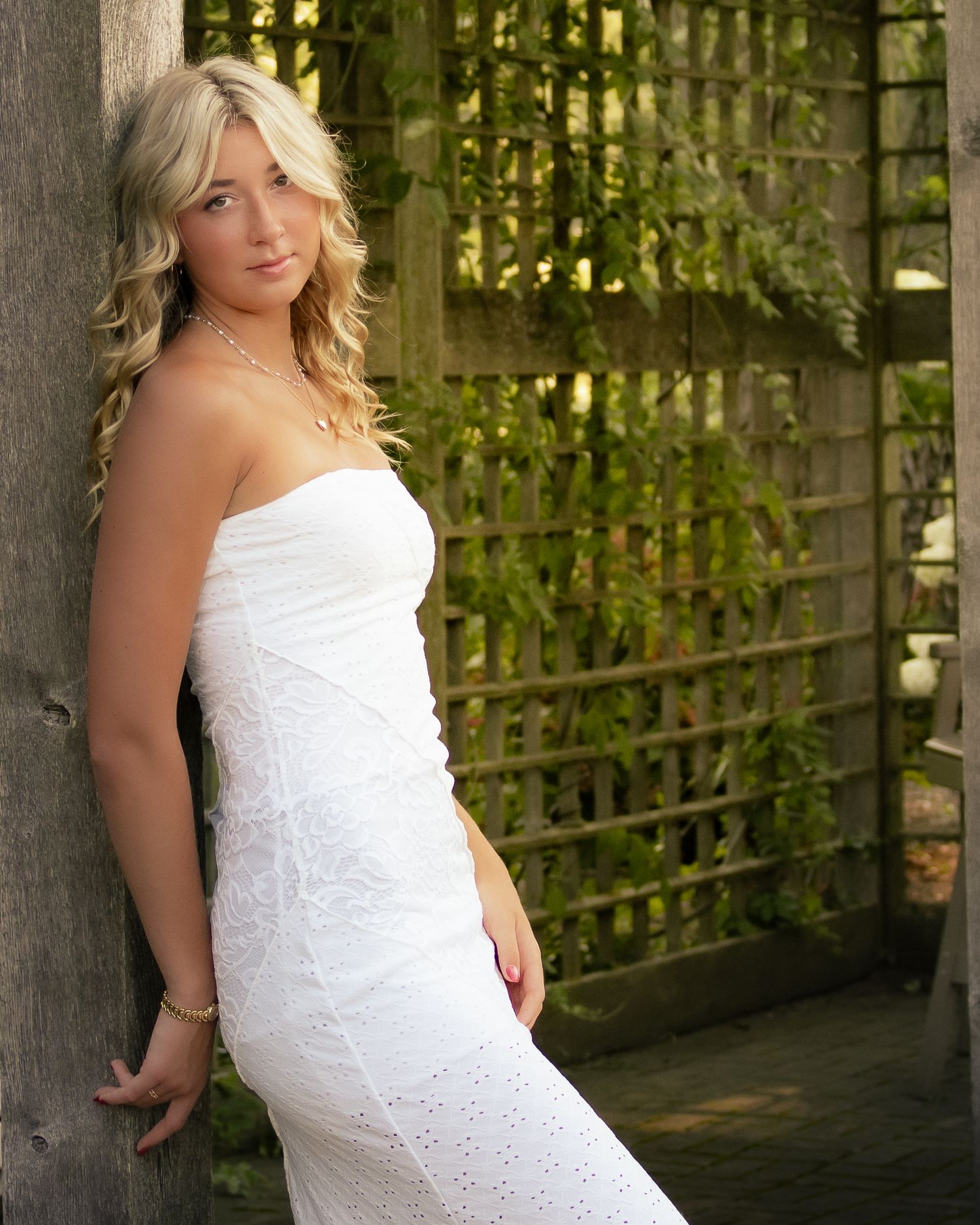 Person in a white eyelet lace dress poses outdoors against a wooden trellis backdrop in natural lighting.