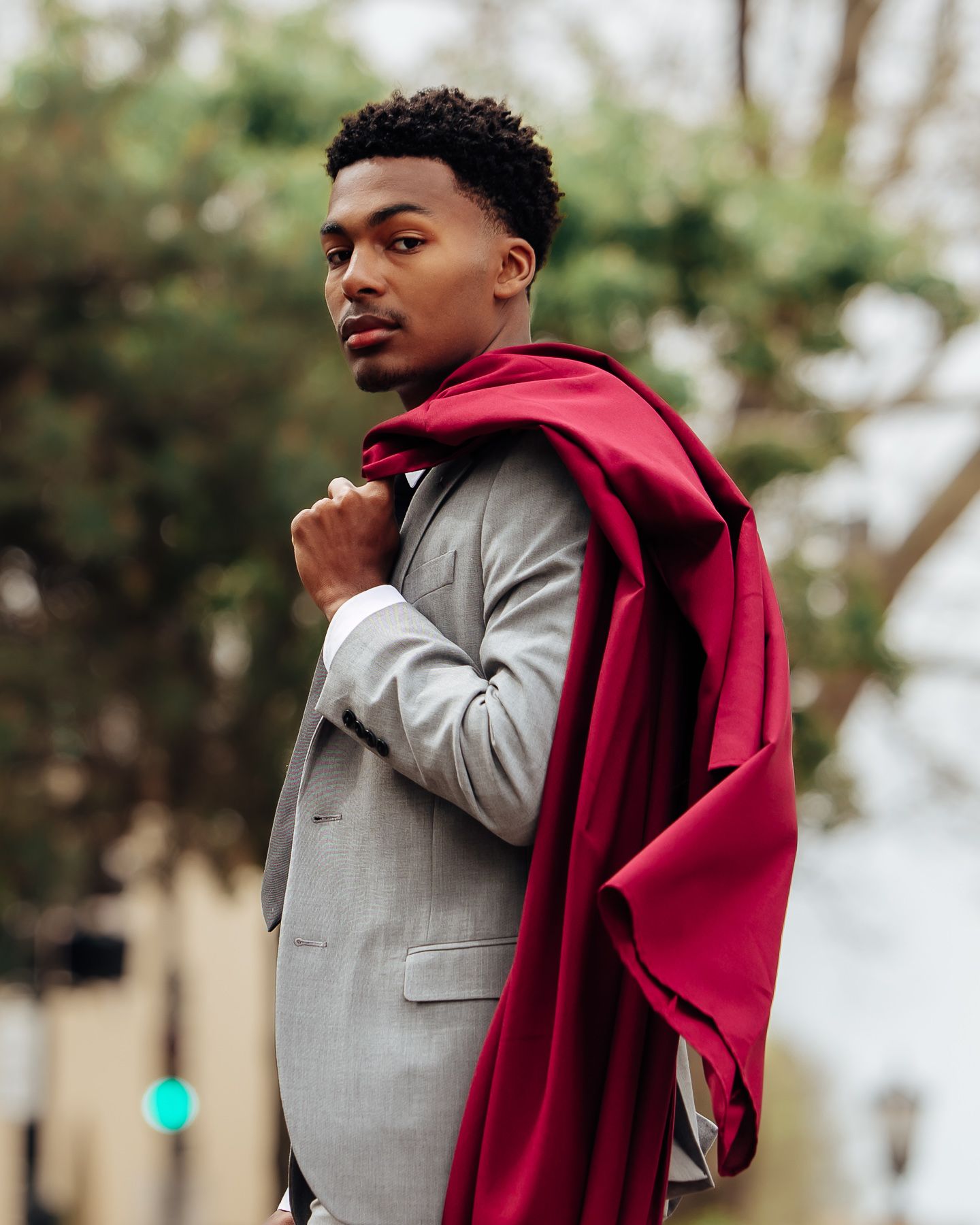 A young man in a gray suit drapes a red coat over his shoulder outdoors.