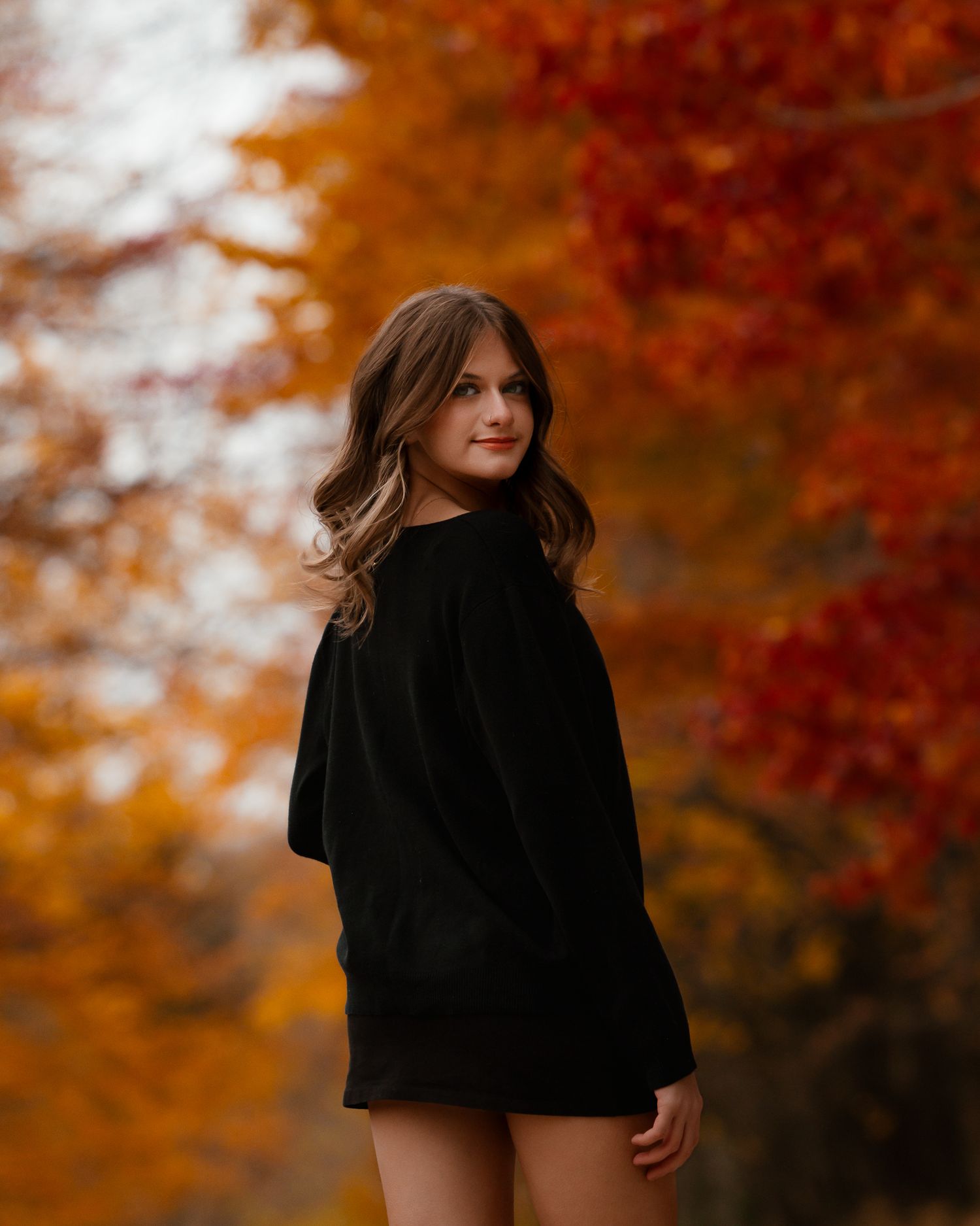 A figure in a black coat poses against a backdrop of vibrant autumn foliage with warm orange tones.