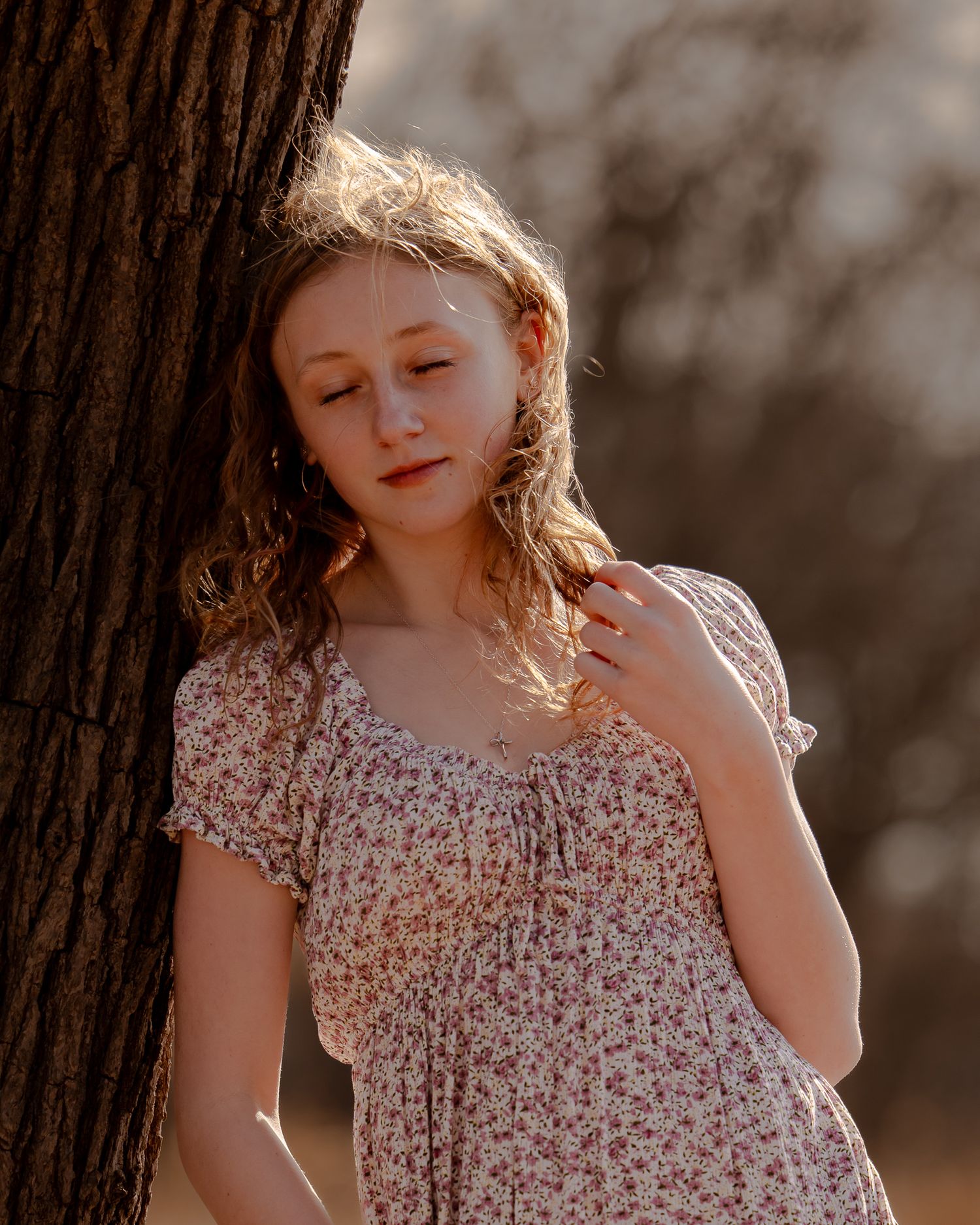 Young woman in floral dress leaning against tree in golden sunlight outdoors.