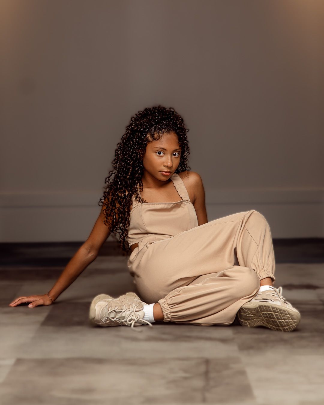 Young Black woman with curly hair sits gracefully on floor wearing a beige jumpsuit and white sneakers.