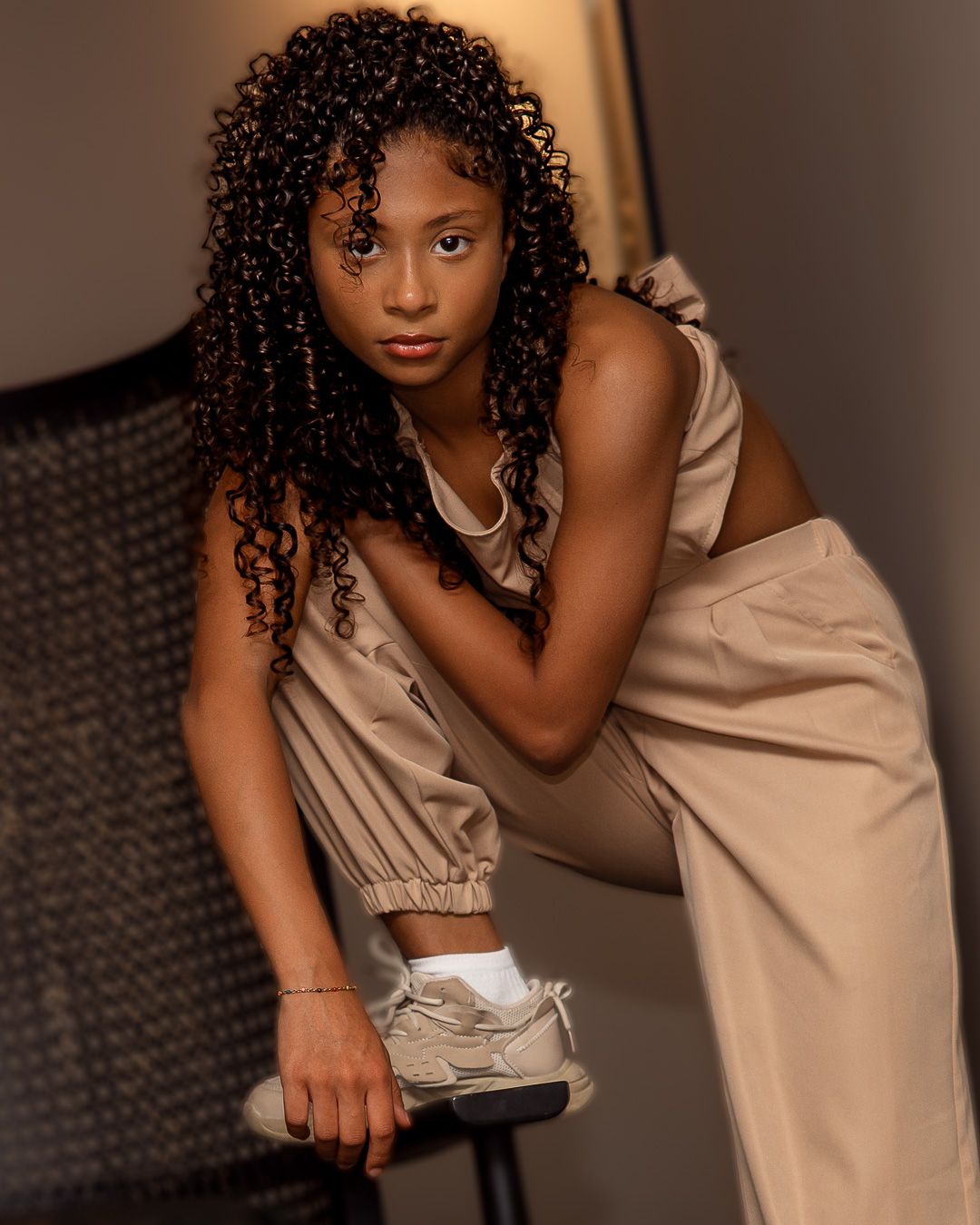 Young woman with curly hair poses confidently in beige outfit with silver sneakers in a studio setting.