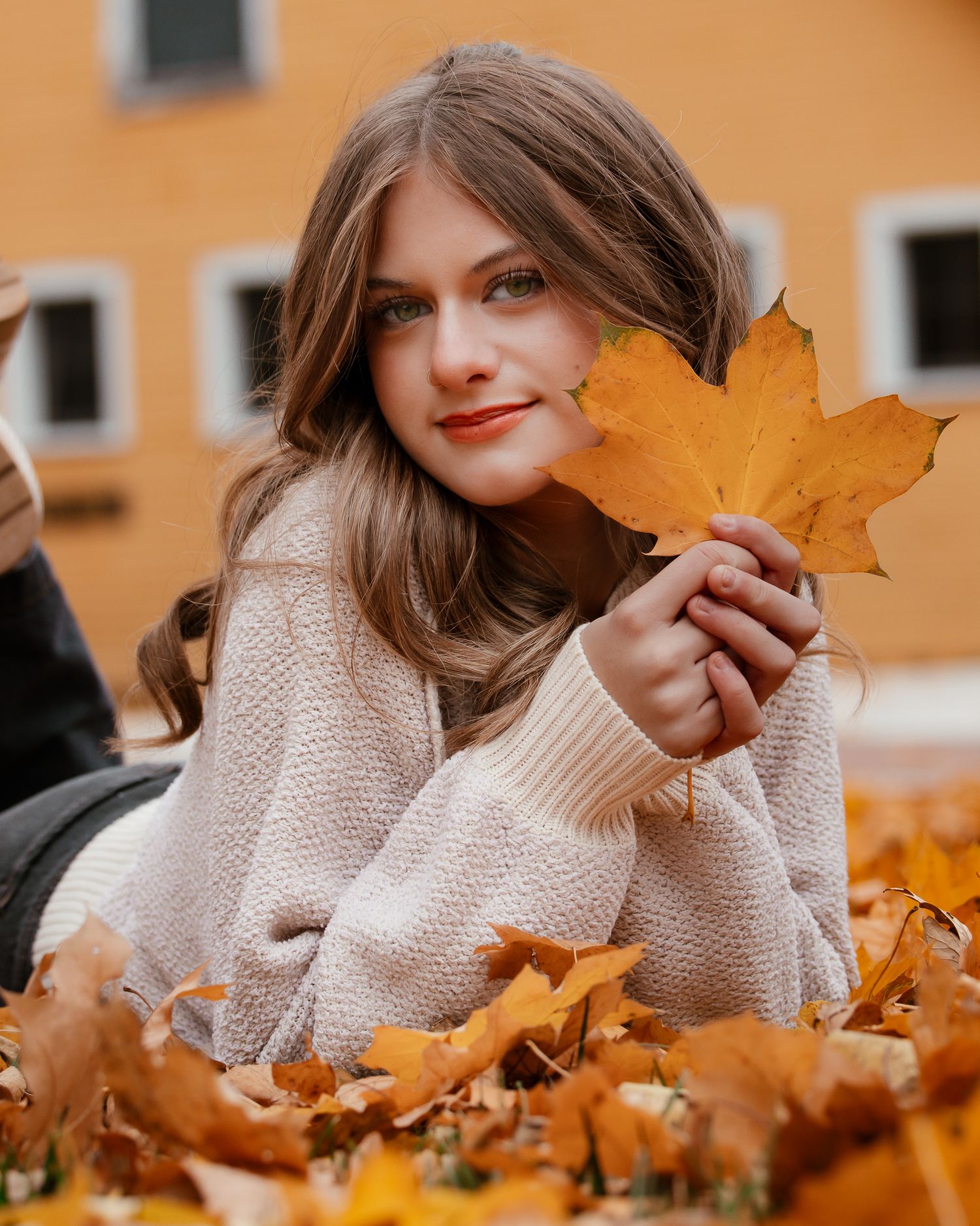 Person in cozy beige sweater lying among autumn leaves holding up a large orange maple leaf against orange building.