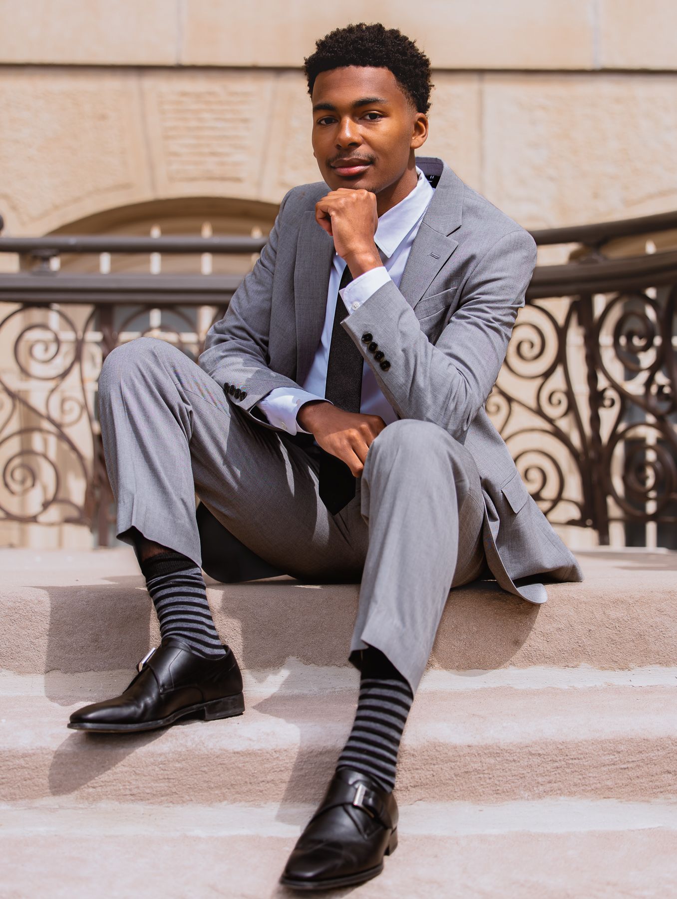 Young man in gray suit with striped socks and black dress shoes sitting on steps in a relaxed pose.
