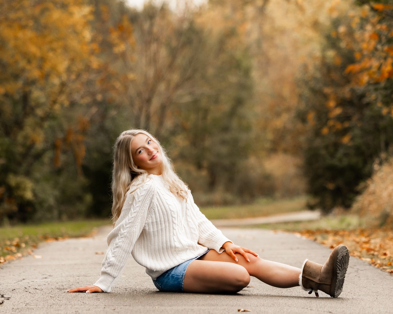 Person in white sweater and shorts lounging on autumn path with fallen leaves.