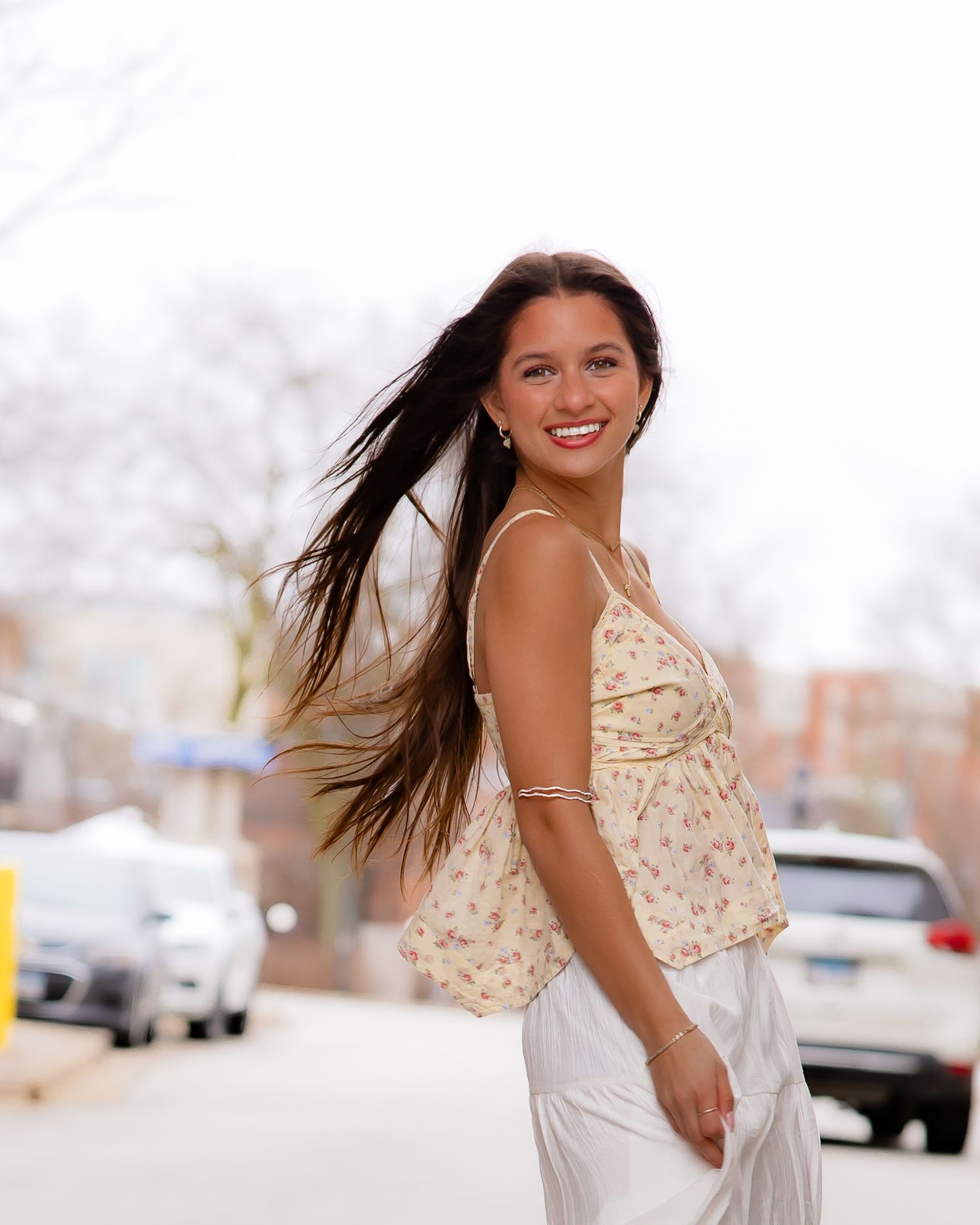 Smiling young woman with long dark hair wearing a floral top and white pants on a city street.