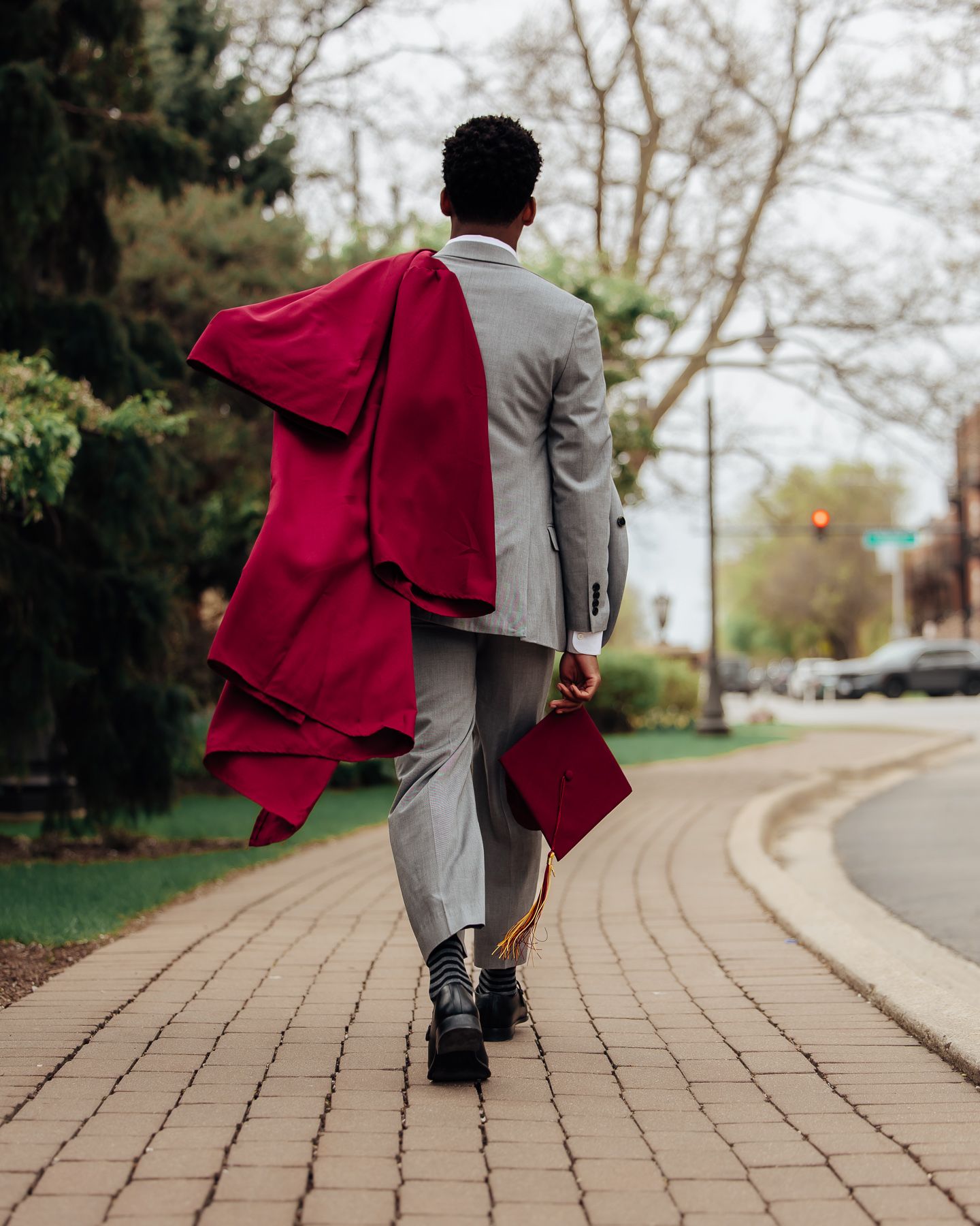 Graduate in gray suit with flowing red gown and cap walks down brick path, trees in background.