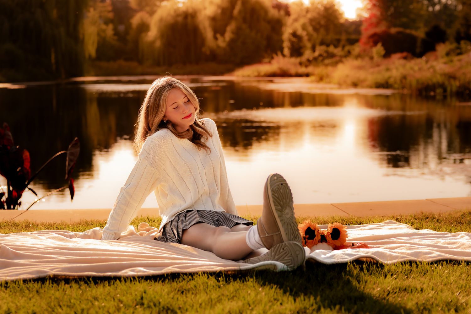 Young woman relaxing on a blanket by a sunlit lake in autumn, wearing a white sweater and denim skirt.