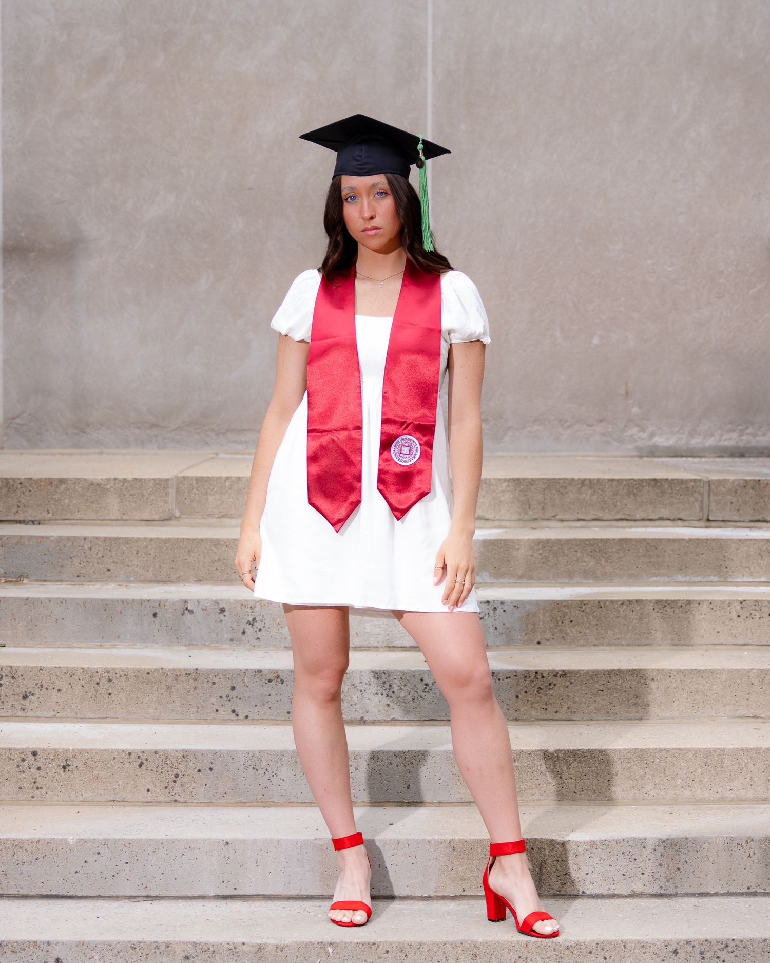 Graduate in white dress and red stole poses on concrete steps wearing graduation cap and red heels.