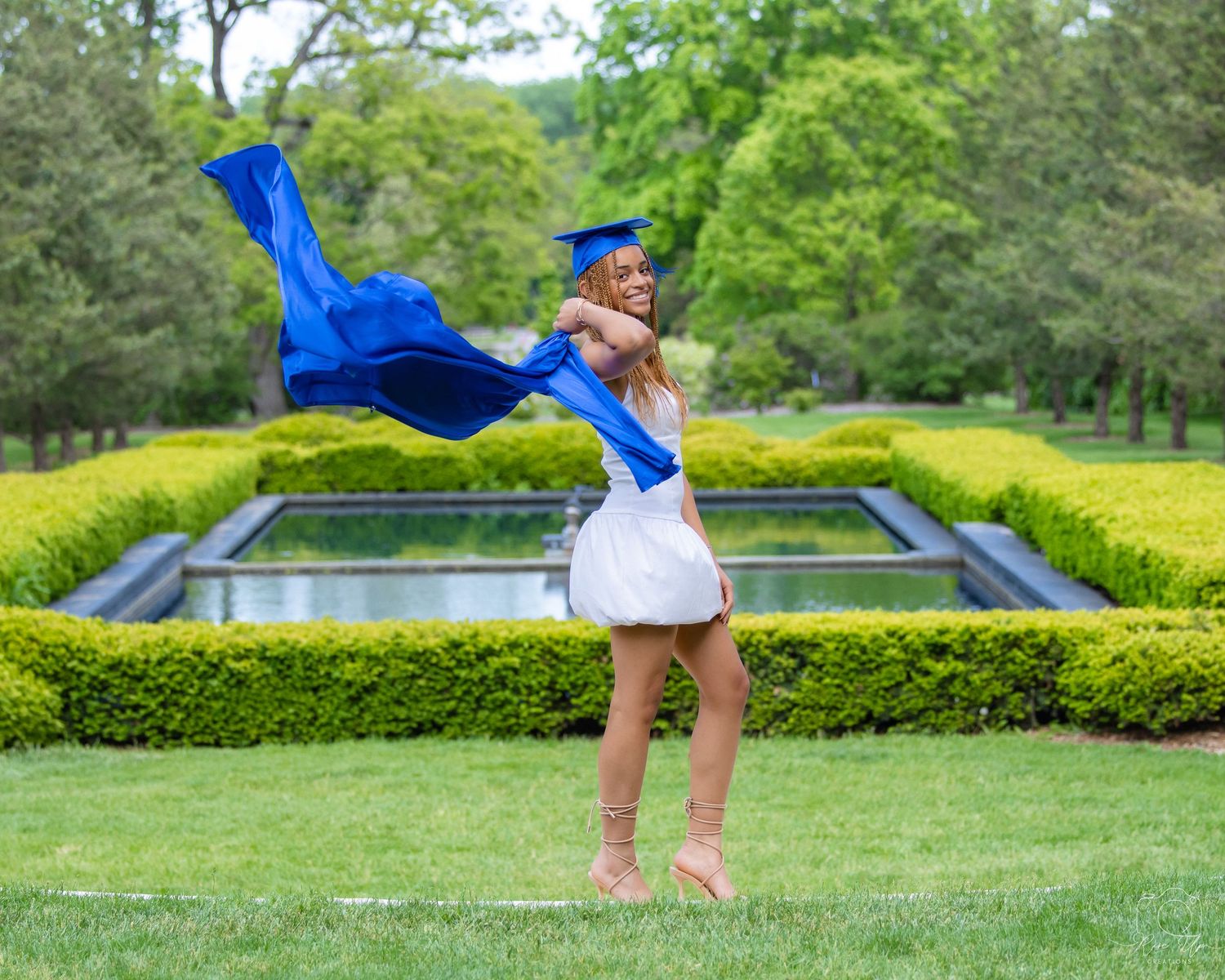 Graduate celebrates by tossing blue graduation gown in formal garden setting.