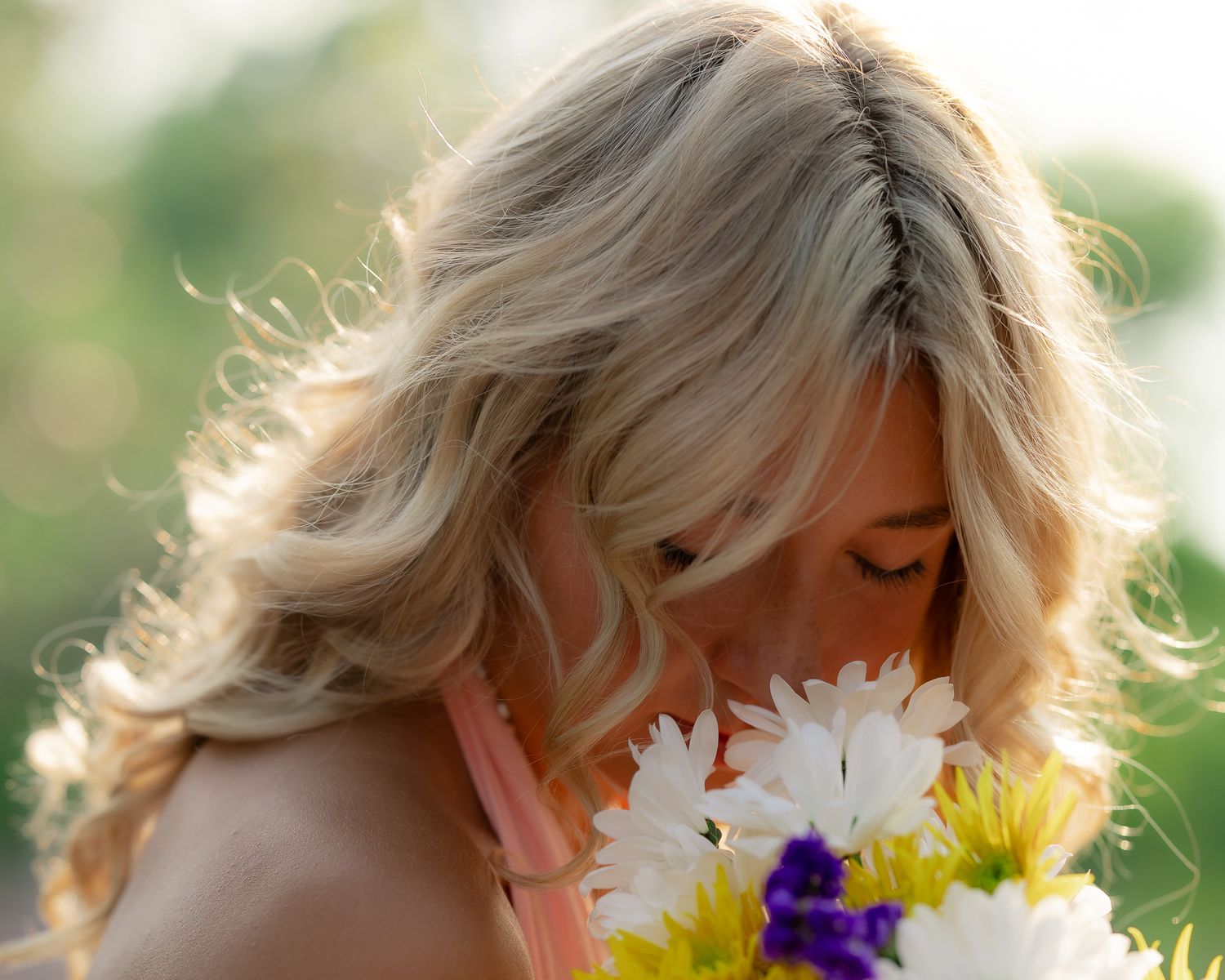 Blonde hair catching sunlight while leaning forward to smell colorful spring flowers in a bouquet.