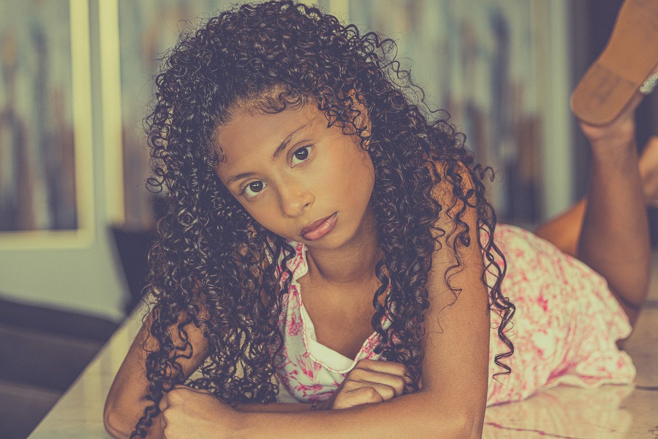 Young girl with long curly dark hair lying down, looking thoughtfully at camera in warm toned photo.