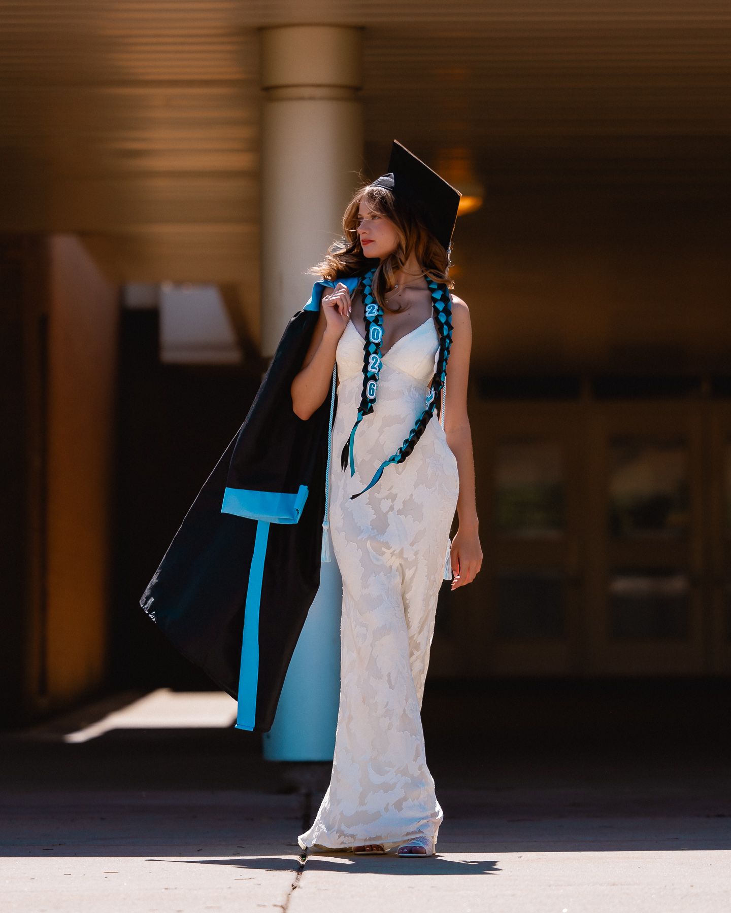 A young woman in a white lace gown and graduation cap holds her black and blue academic robe outdoors.