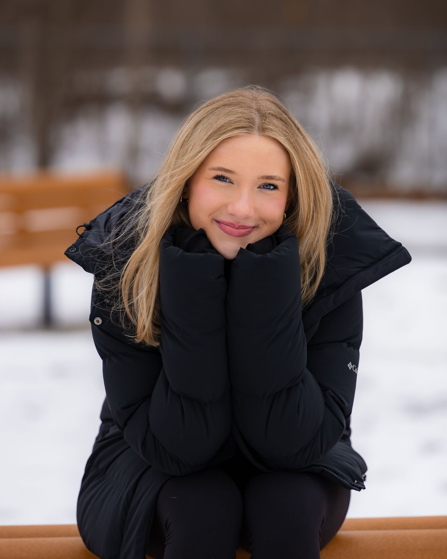 Smiling blonde girl in black puffer jacket sitting on snowy bench outdoors in winter park.