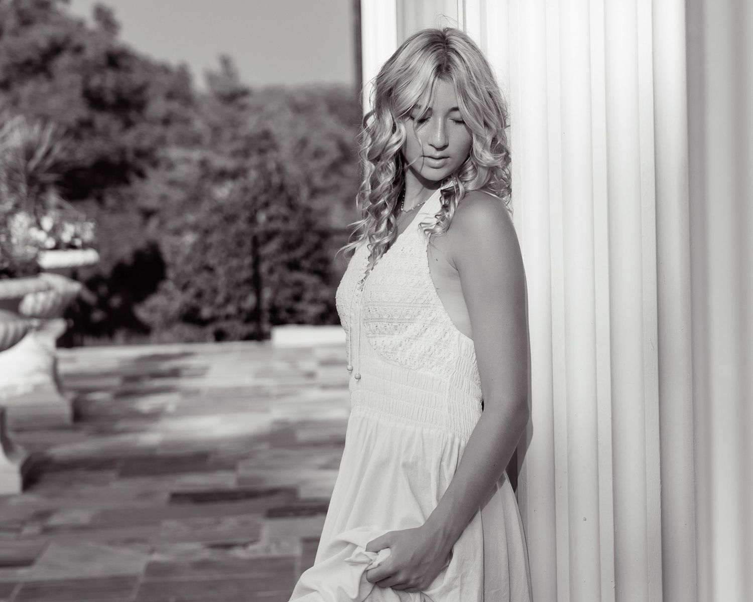 Black and white portrait of a woman in white dress leaning against a column outdoors.