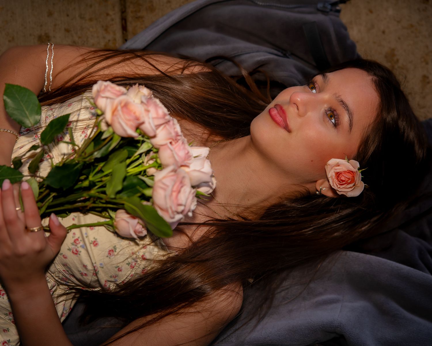 Young woman lying down holding pink roses bouquet with a rose in her hair, wearing floral dress.