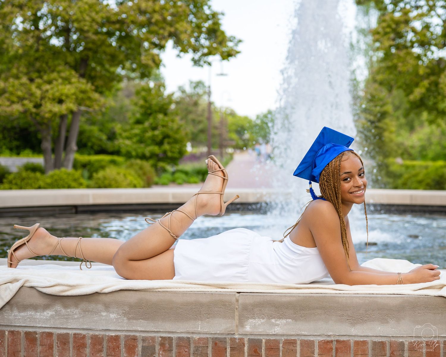 Graduate in white dress lying on brick ledge near fountain on sunny campus day.