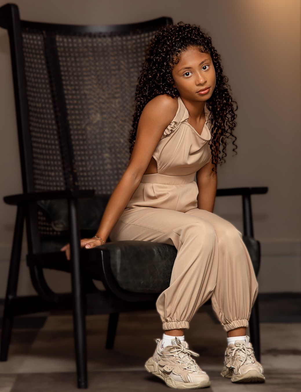 Fashion portrait of model in beige jumpsuit and sneakers posing on chair in studio with moody lighting.