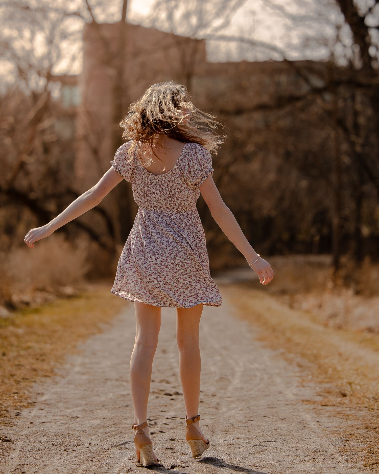 A woman in a floral mini dress twirls on a dirt path surrounded by bare trees in warm golden hour light.