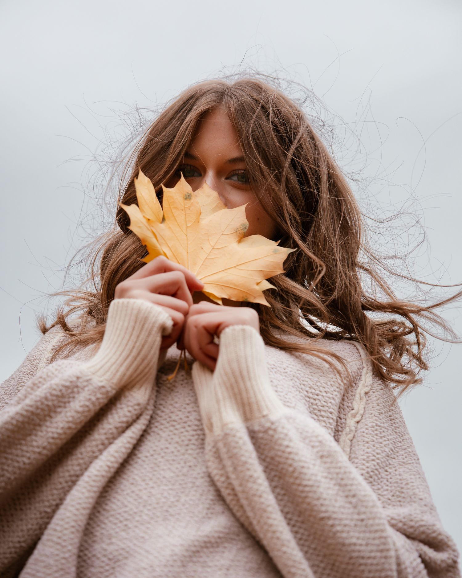 A person in a beige sweater playfully holds an autumn maple leaf in front of their face in a series of photos.