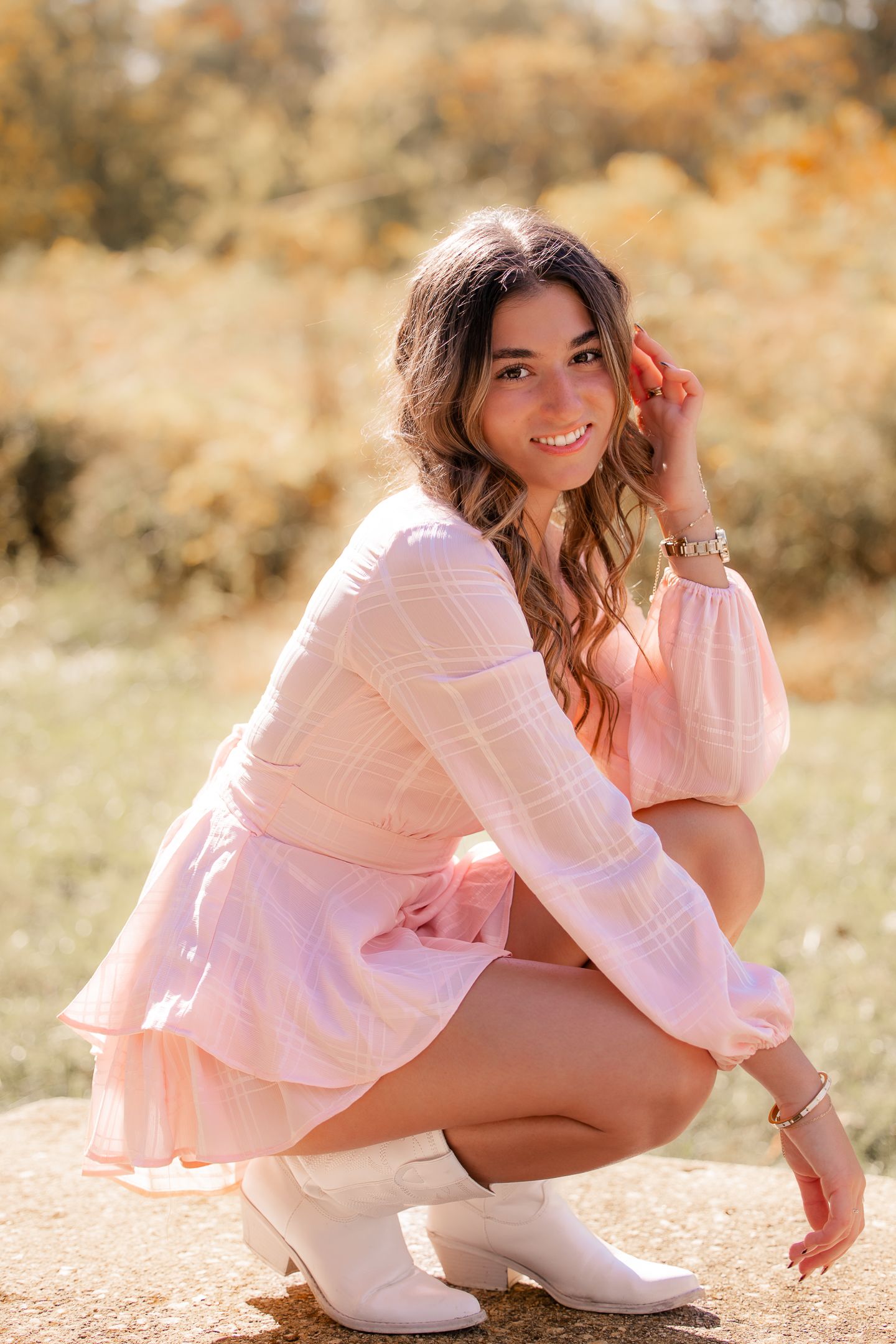 A person in a pink sweater and white boots poses outdoors during golden hour in a natural autumn setting.