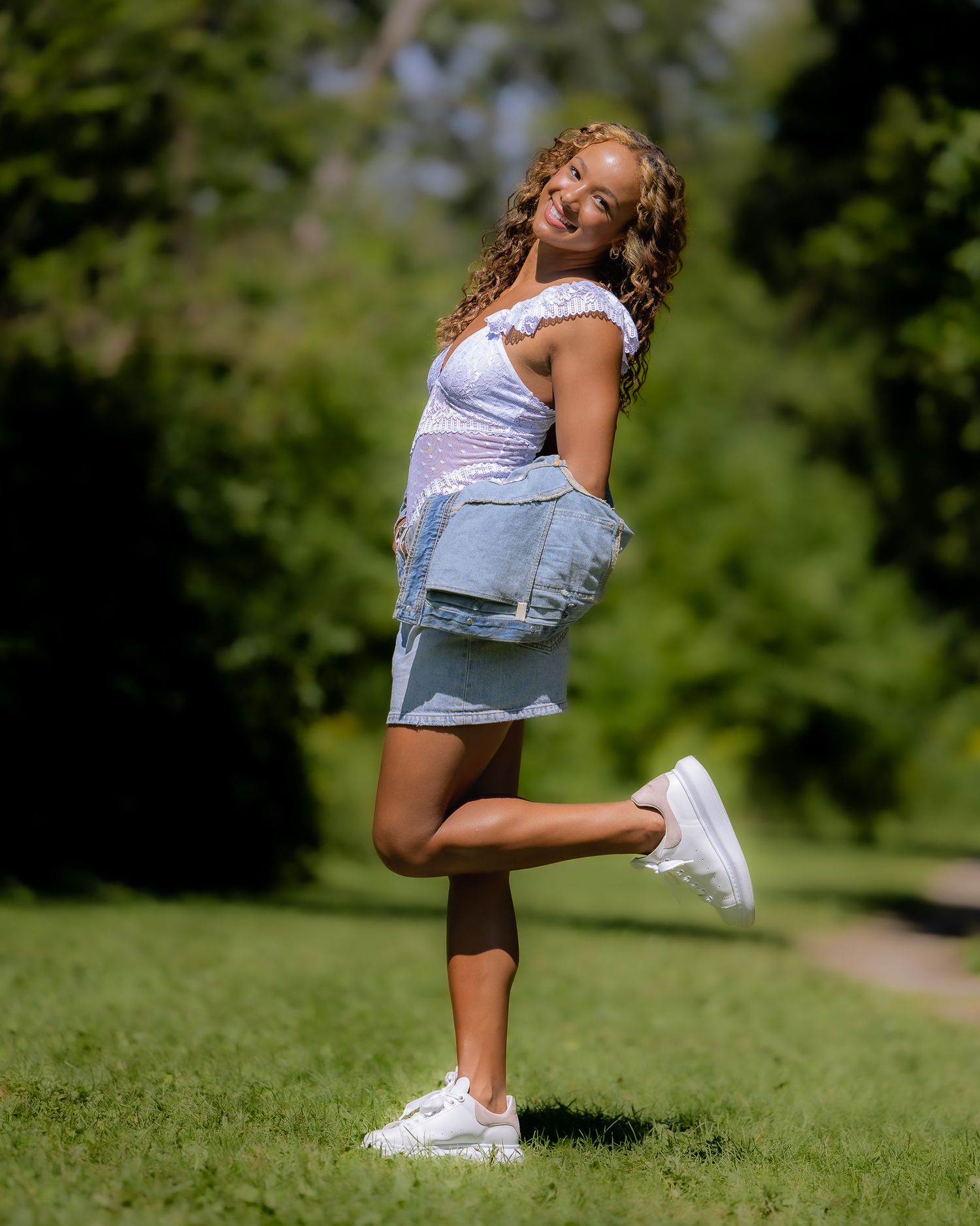 Young person in white top and denim shorts poses playfully on one leg against green outdoor background.
