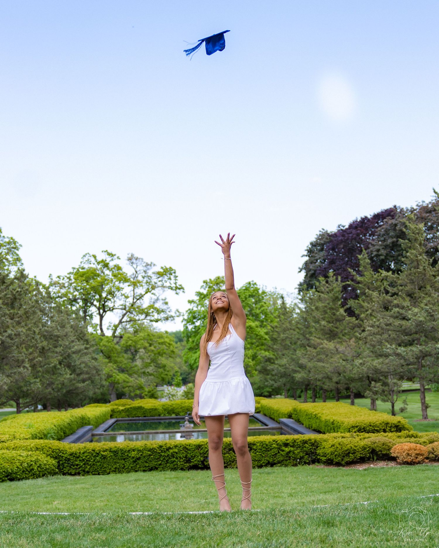 Graduate in white dress tosses blue graduation cap in manicured garden.