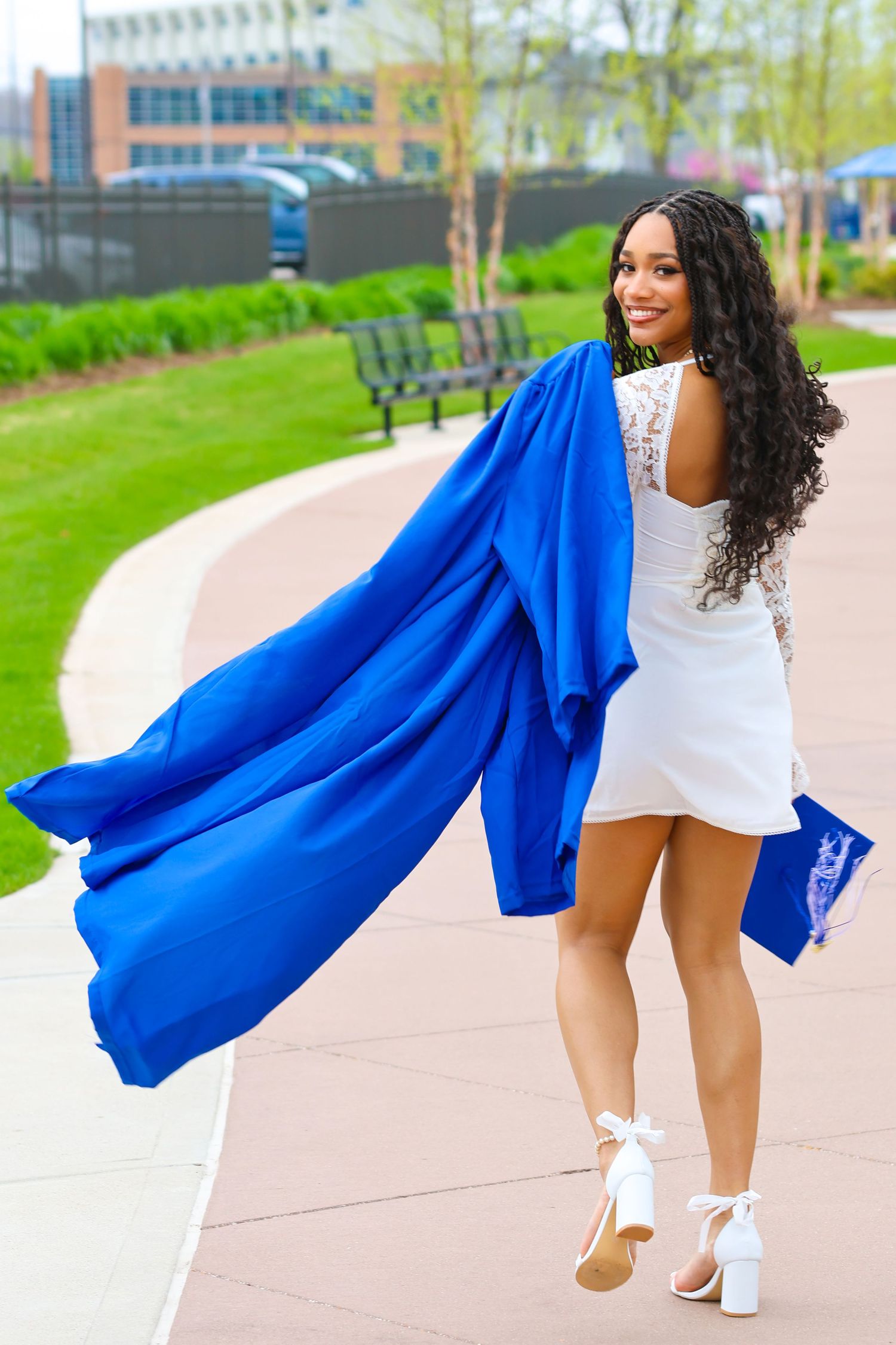 Graduate in white dress twirls with flowing blue graduation gown on campus walkway.