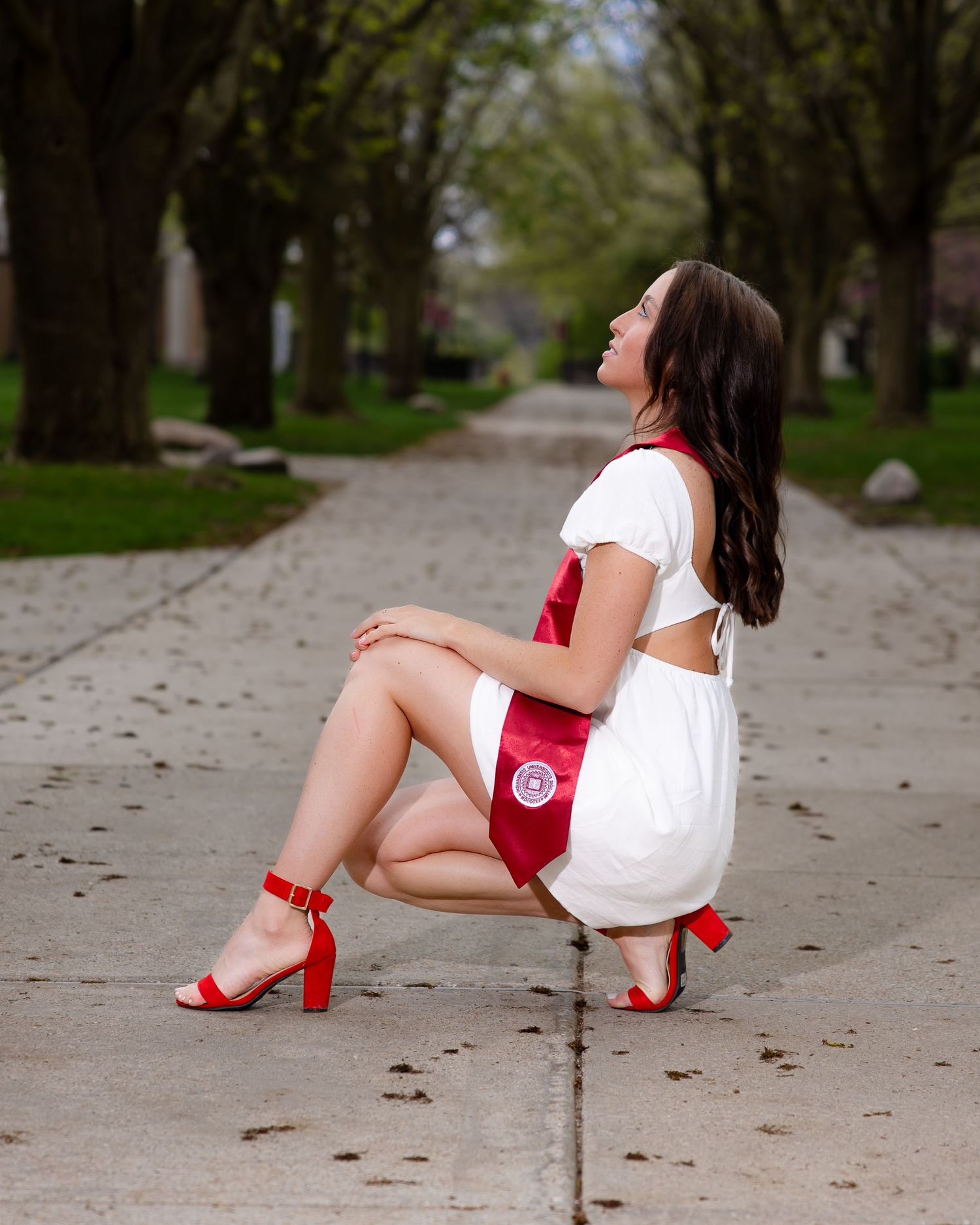 Woman in white dress and red heels poses on a tree-lined path during a graduation photoshoot.