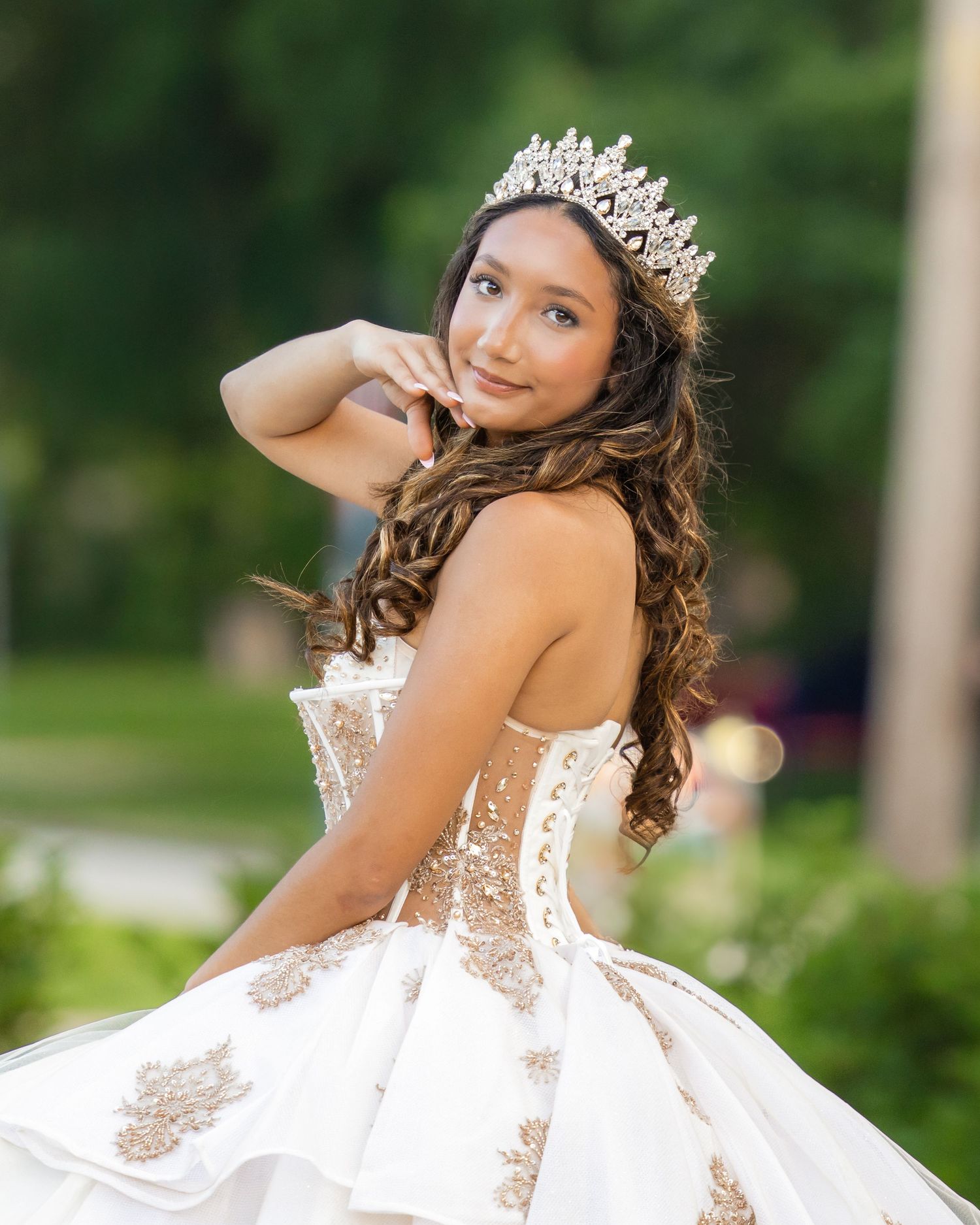 A young person in a white embroidered quinceanera dress and crystal crown poses elegantly in an outdoor garden setting.