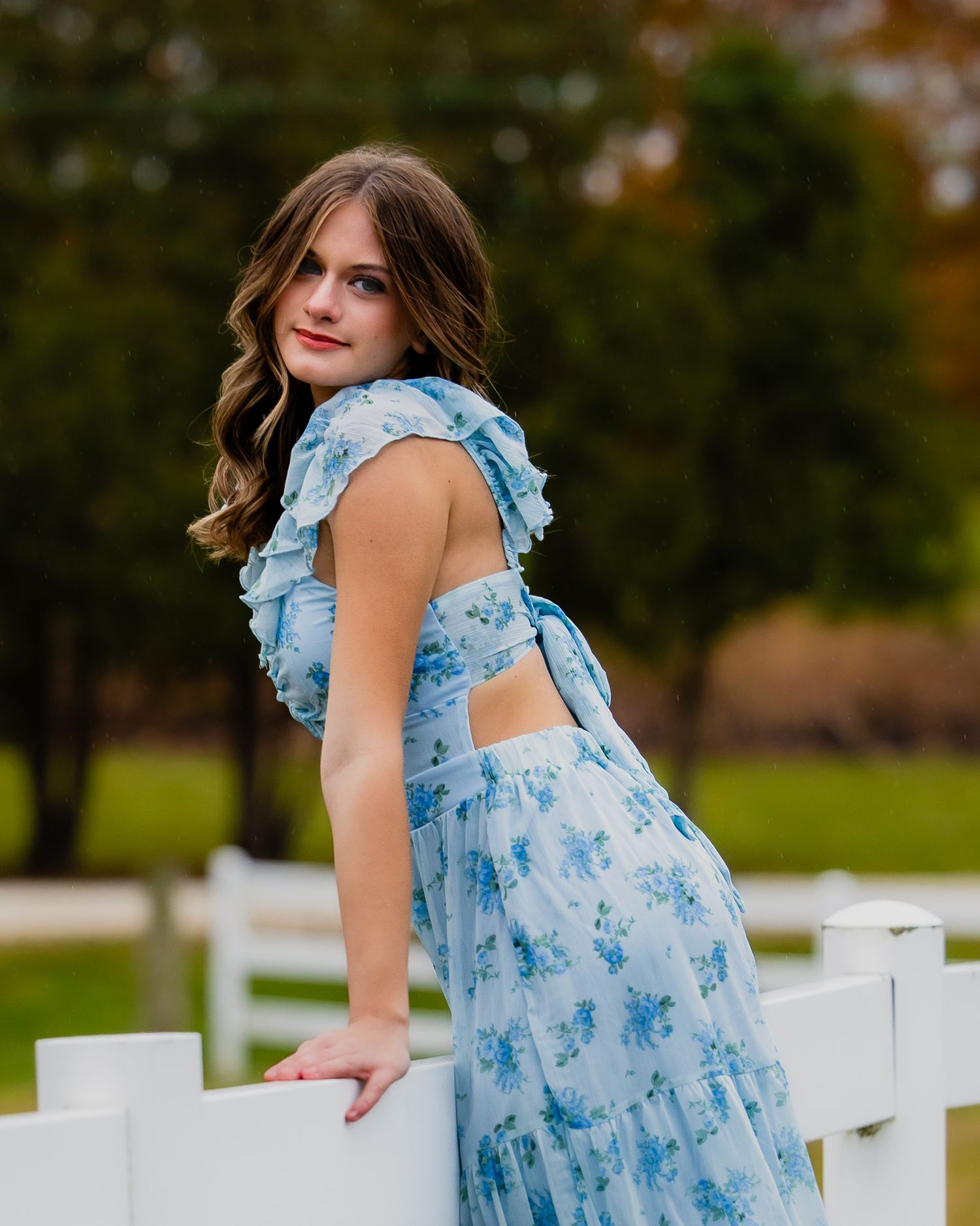 Someone wearing a light blue floral dress poses by white fence posts outdoors.