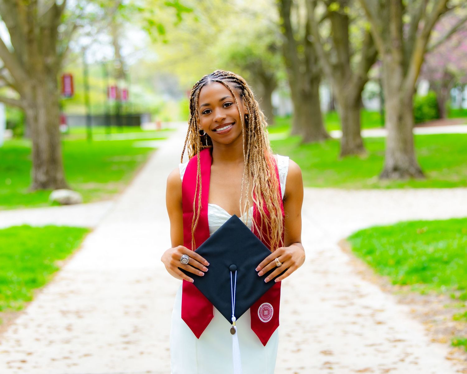 Graduate in red and white graduation regalia poses with cap on tree-lined walkway during spring.