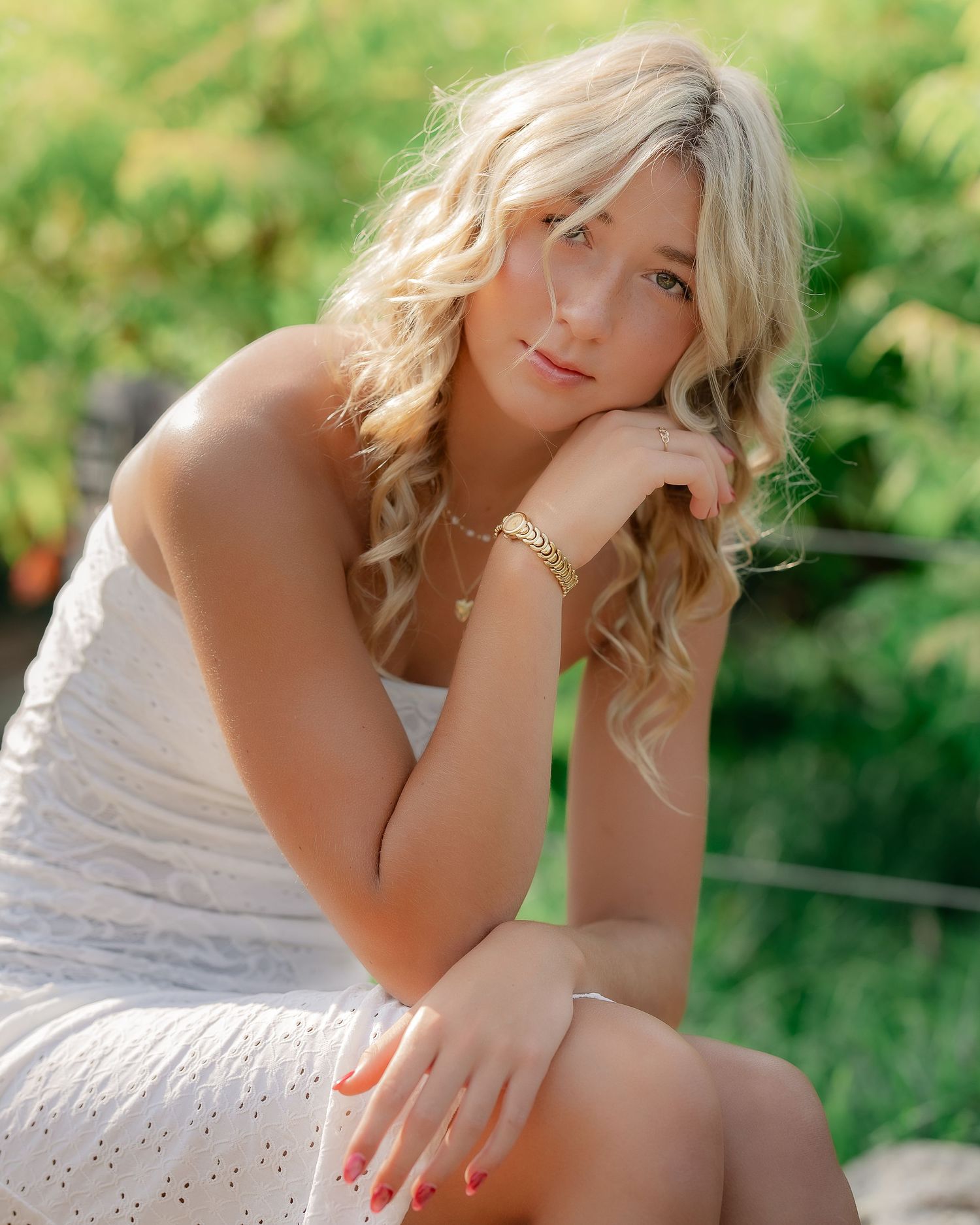 A person in a white sparkly dress poses outdoors with blonde curly hair and a gold bracelet against a green background.