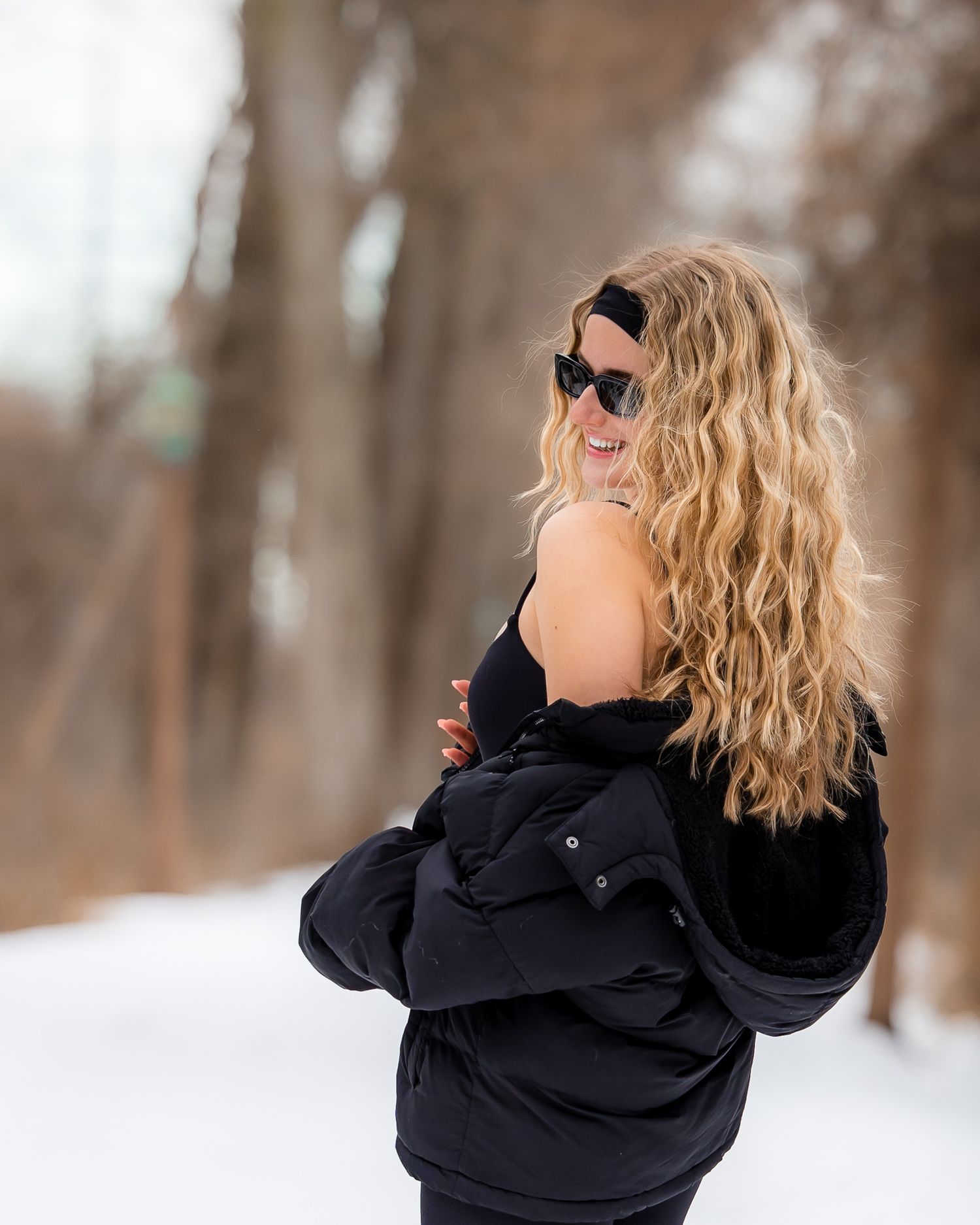 A person with wavy blonde hair wearing a black winter coat poses playfully in a snowy forest setting.