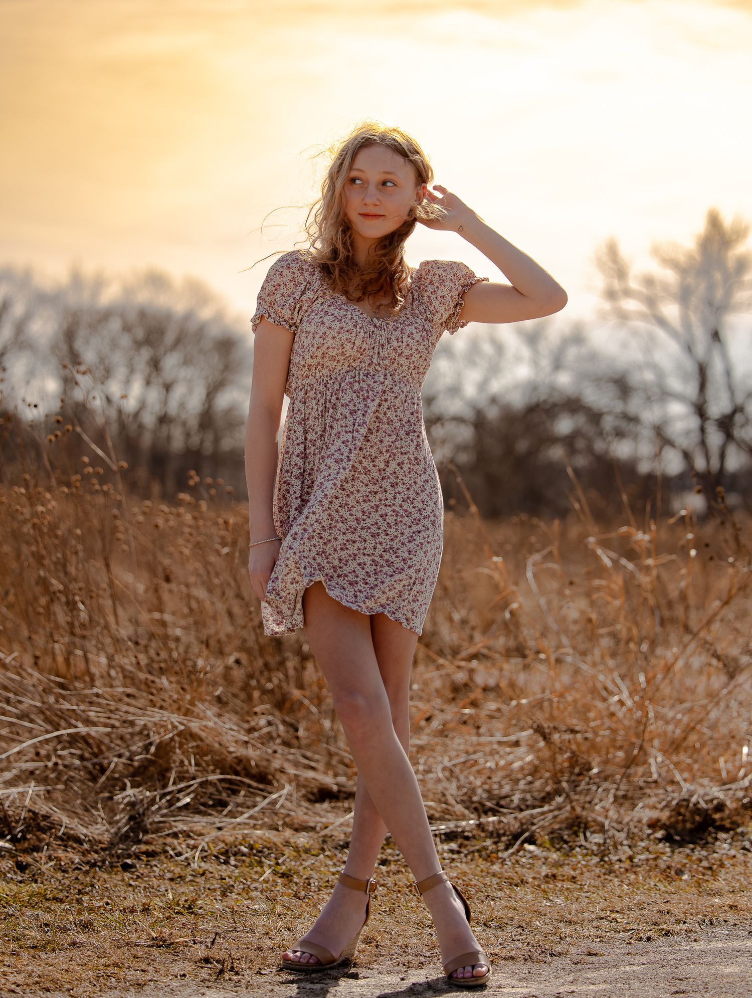 Young woman in floral mini dress posing outdoors at golden sunset with dry grass field background.