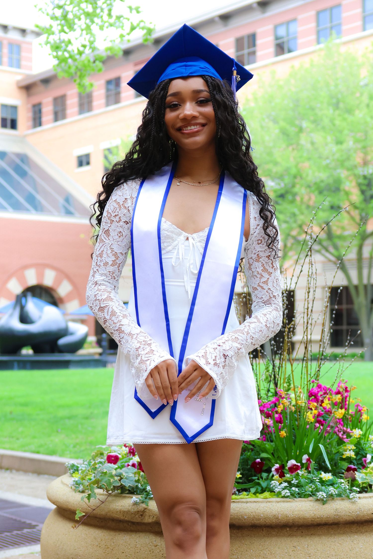 Graduate in white lace dress and blue graduation stole poses near garden with flowers.