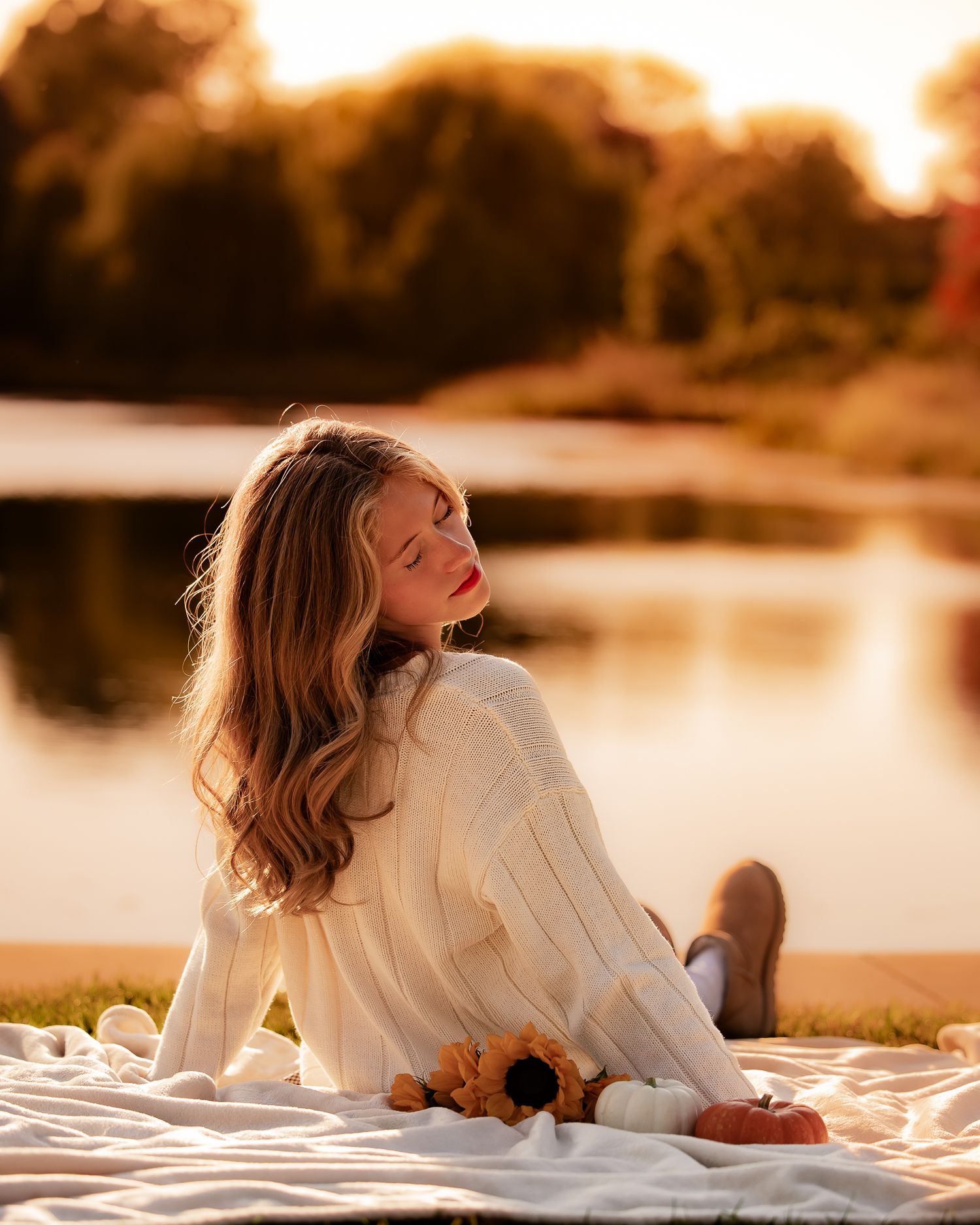 Young woman in cream sweater sitting on blanket by lake at golden hour sunset with sunflowers.