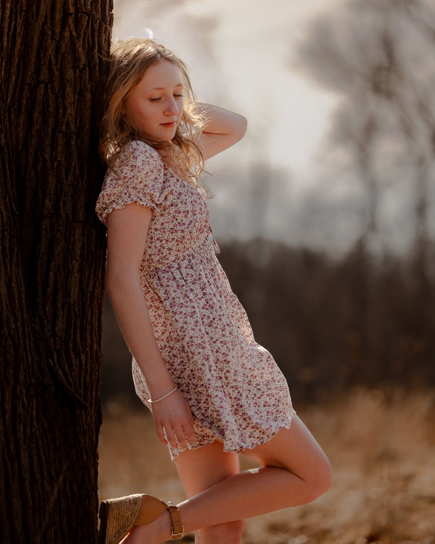 Young blonde woman in floral dress leaning against tree trunk in golden sunlight outdoors.