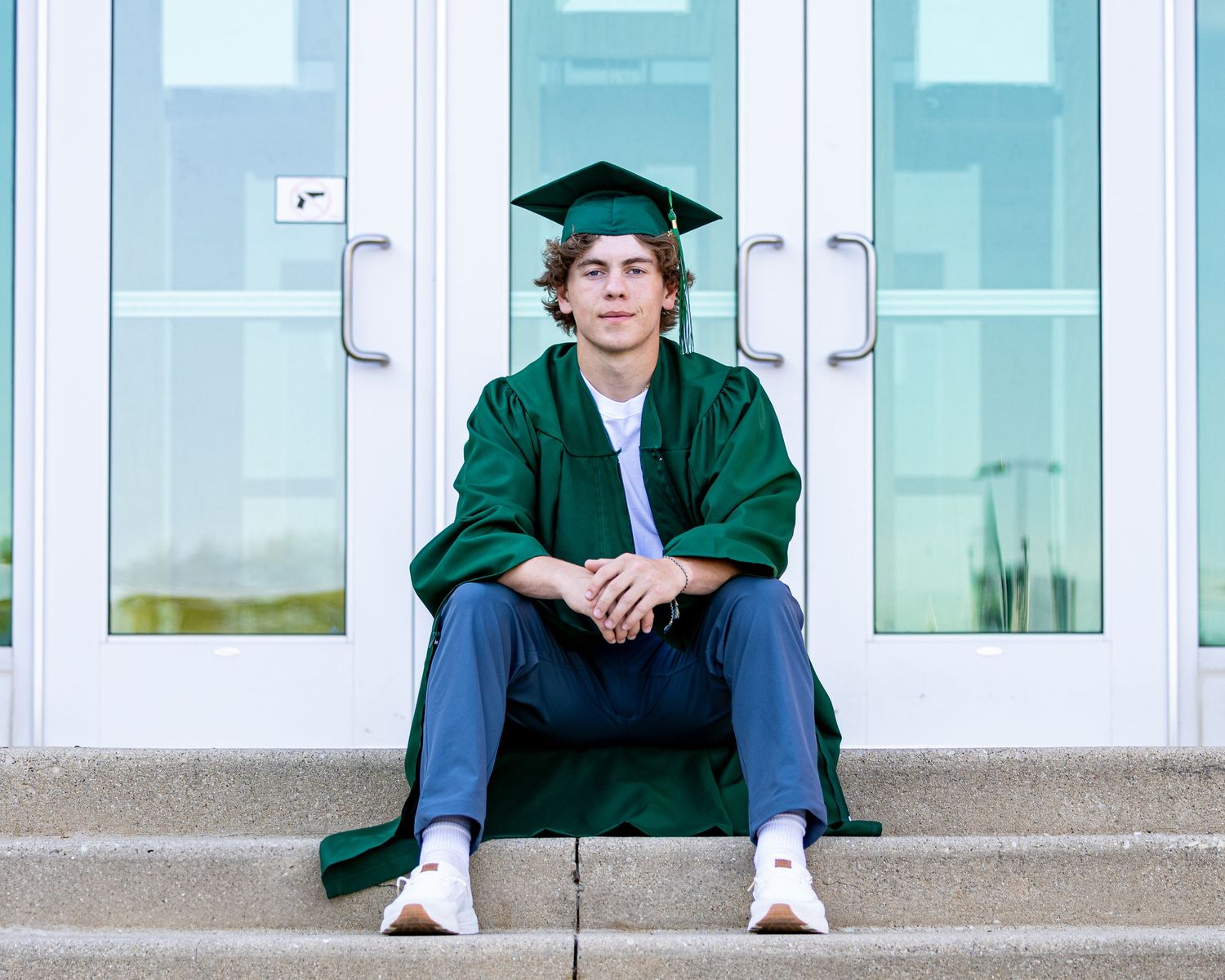Graduate in green cap and gown sits on steps in front of glass doors.