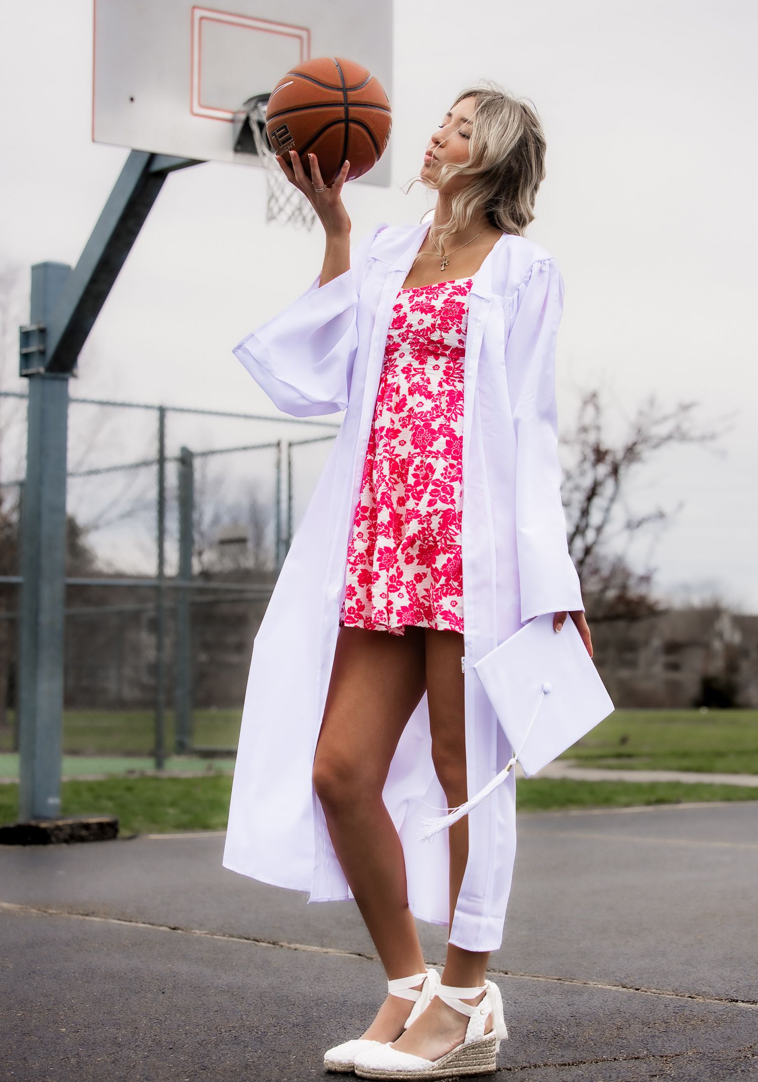 Woman in white graduation gown and floral dress holds basketball on outdoor court with hoop behind her.