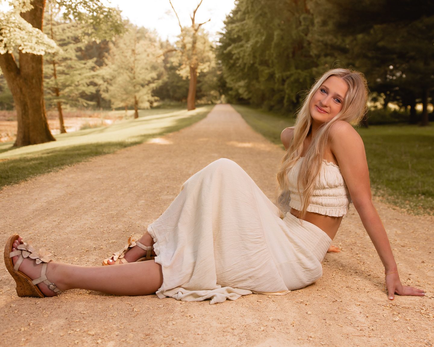 Young blonde woman in white crop top and skirt sitting on a dirt path surrounded by lush green trees.
