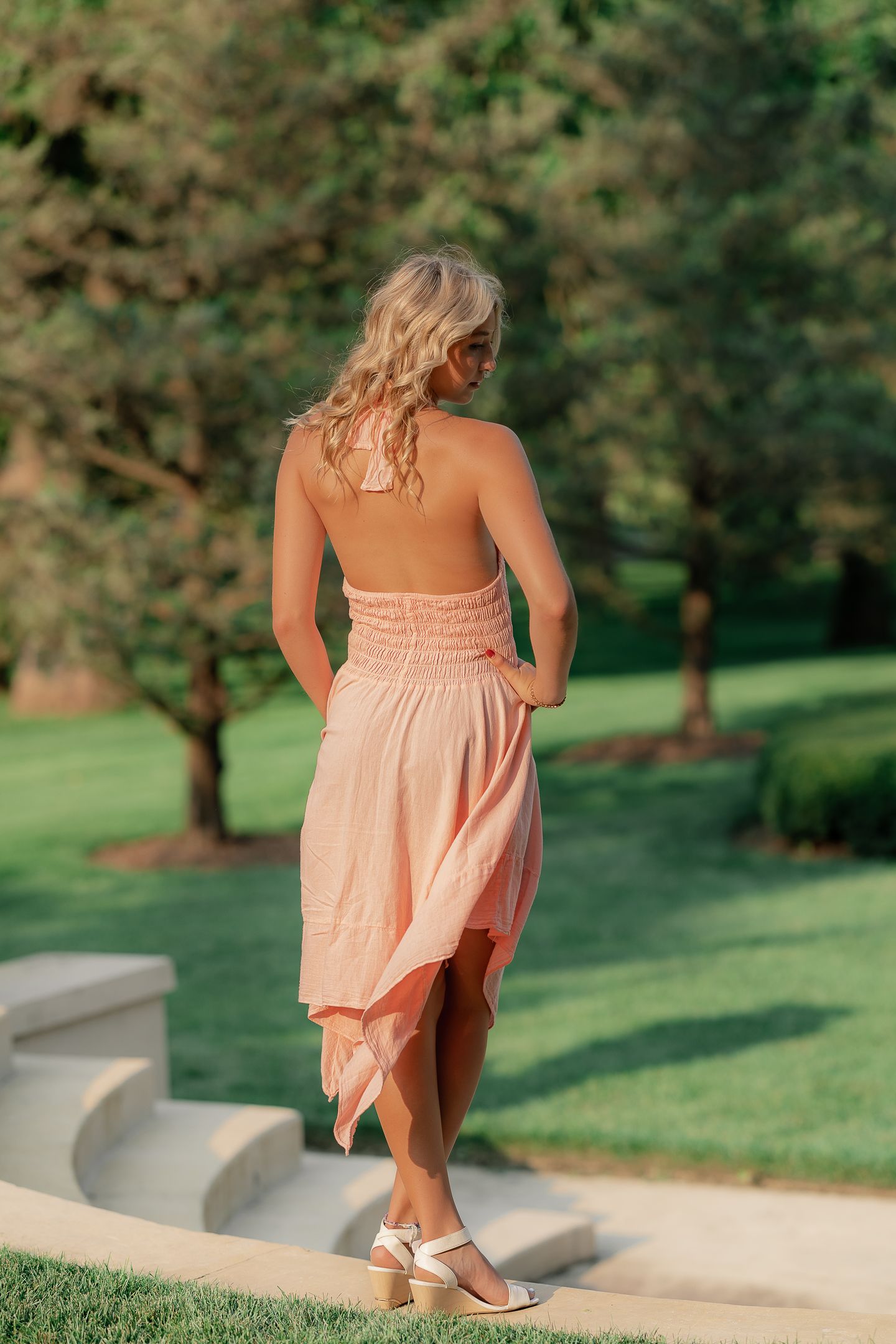 Person in a peach-colored dress sits gracefully on curved concrete wall in a lush garden setting.